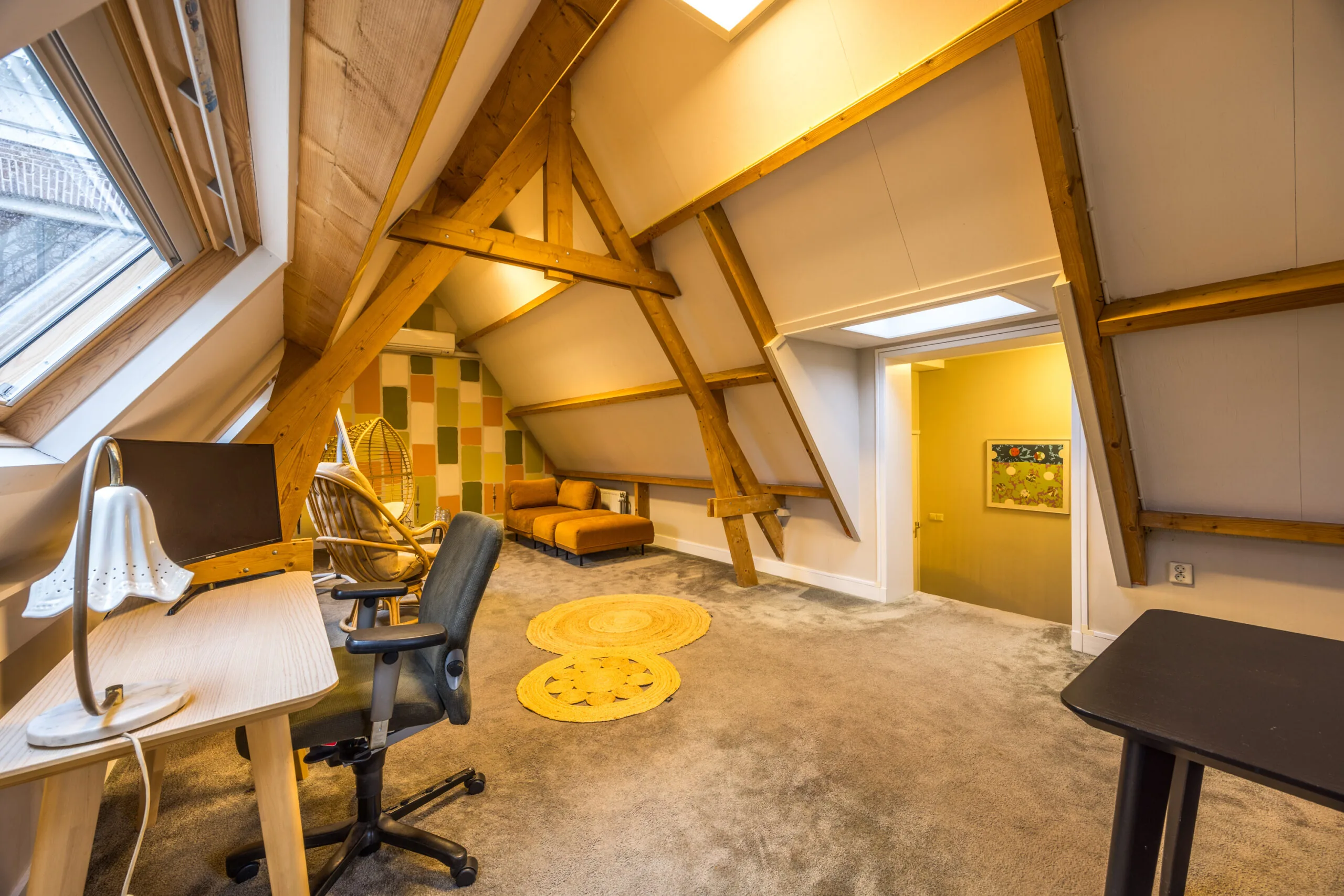 Zolderkamer met werkplek, houten balken, kleurrijke wand en zithoek met rotan schommelstoel.