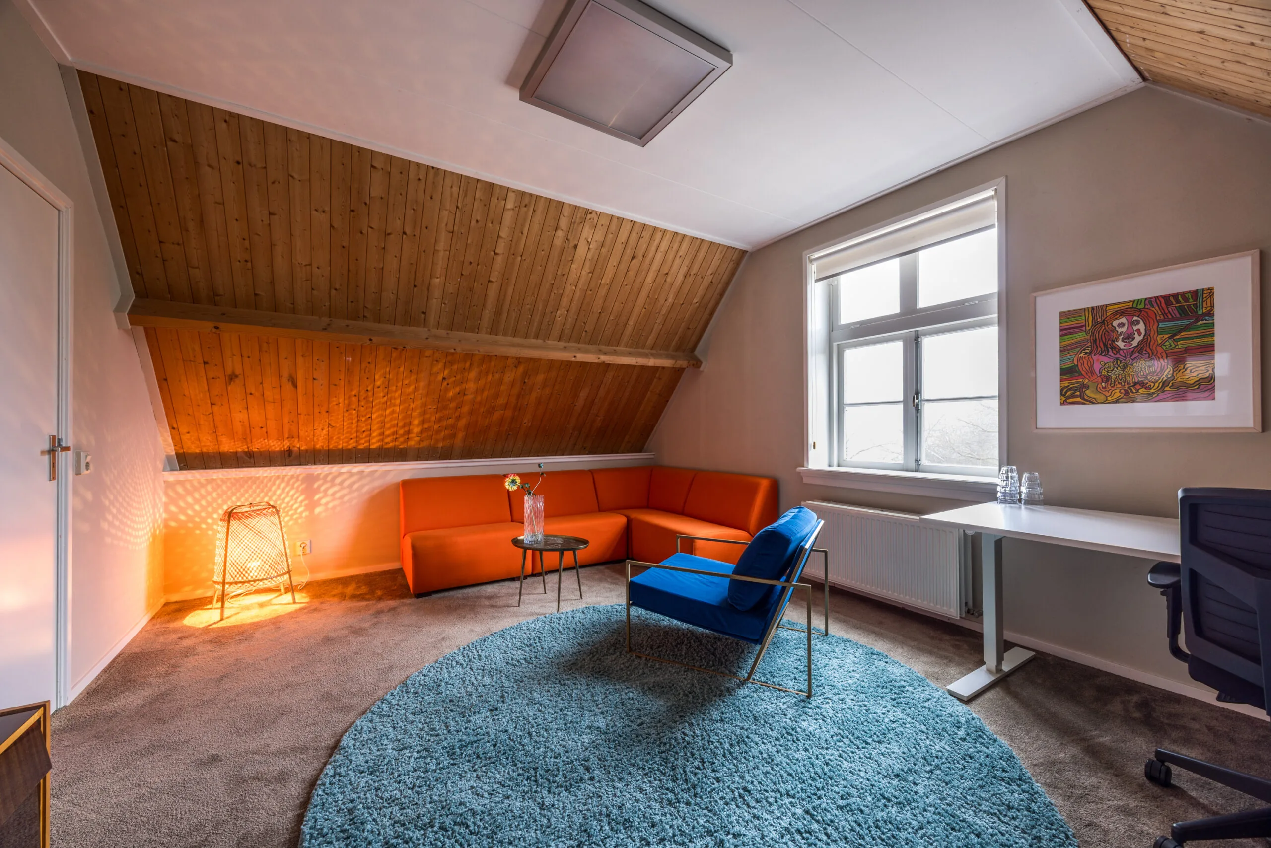 Knusse zolderkamer met een oranje hoekbank, blauwe fauteuil, bureau en zachtblauwe rond vloerkleed onder een schuine houten plafond.