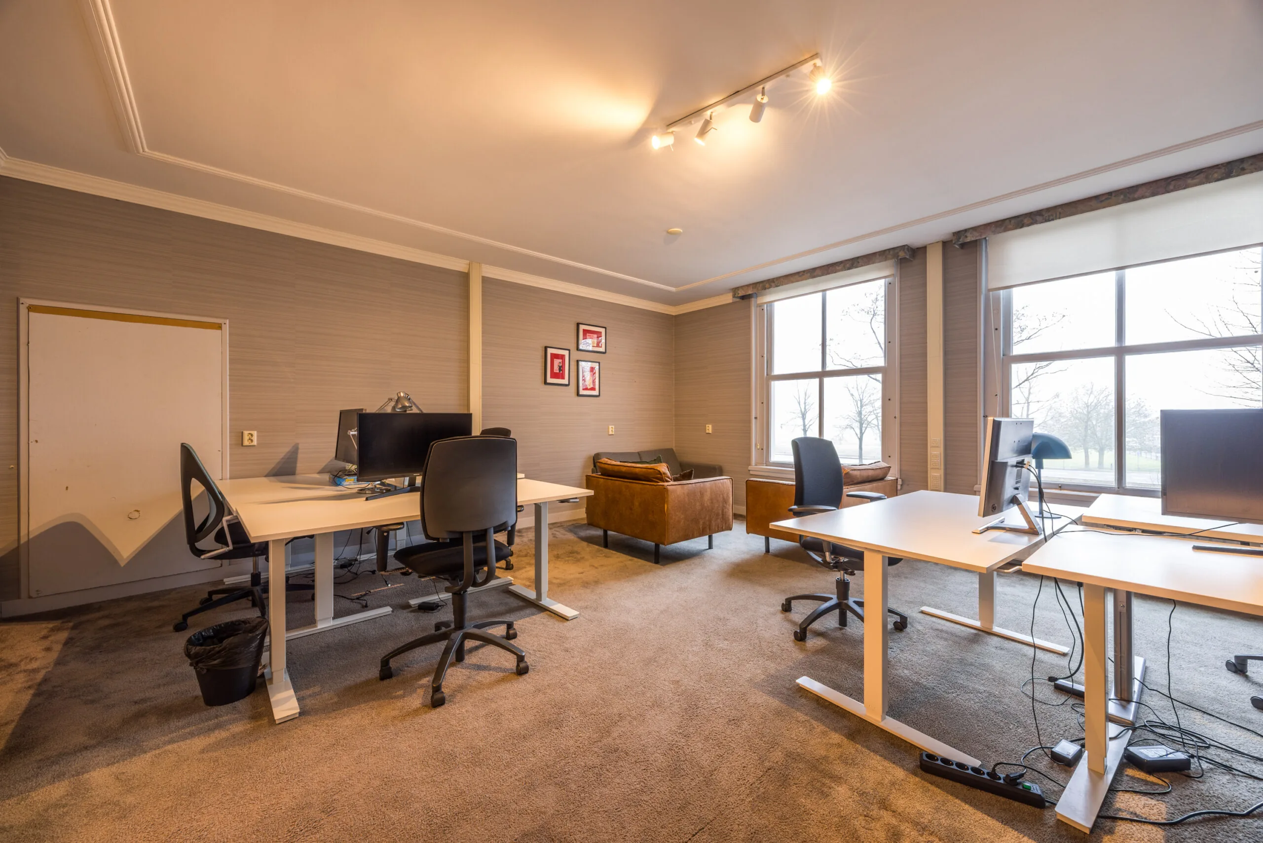 Modern kantoor met bureaus, bureaustoelen, computers en een zithoek bij grote ramen met uitzicht op bomen.