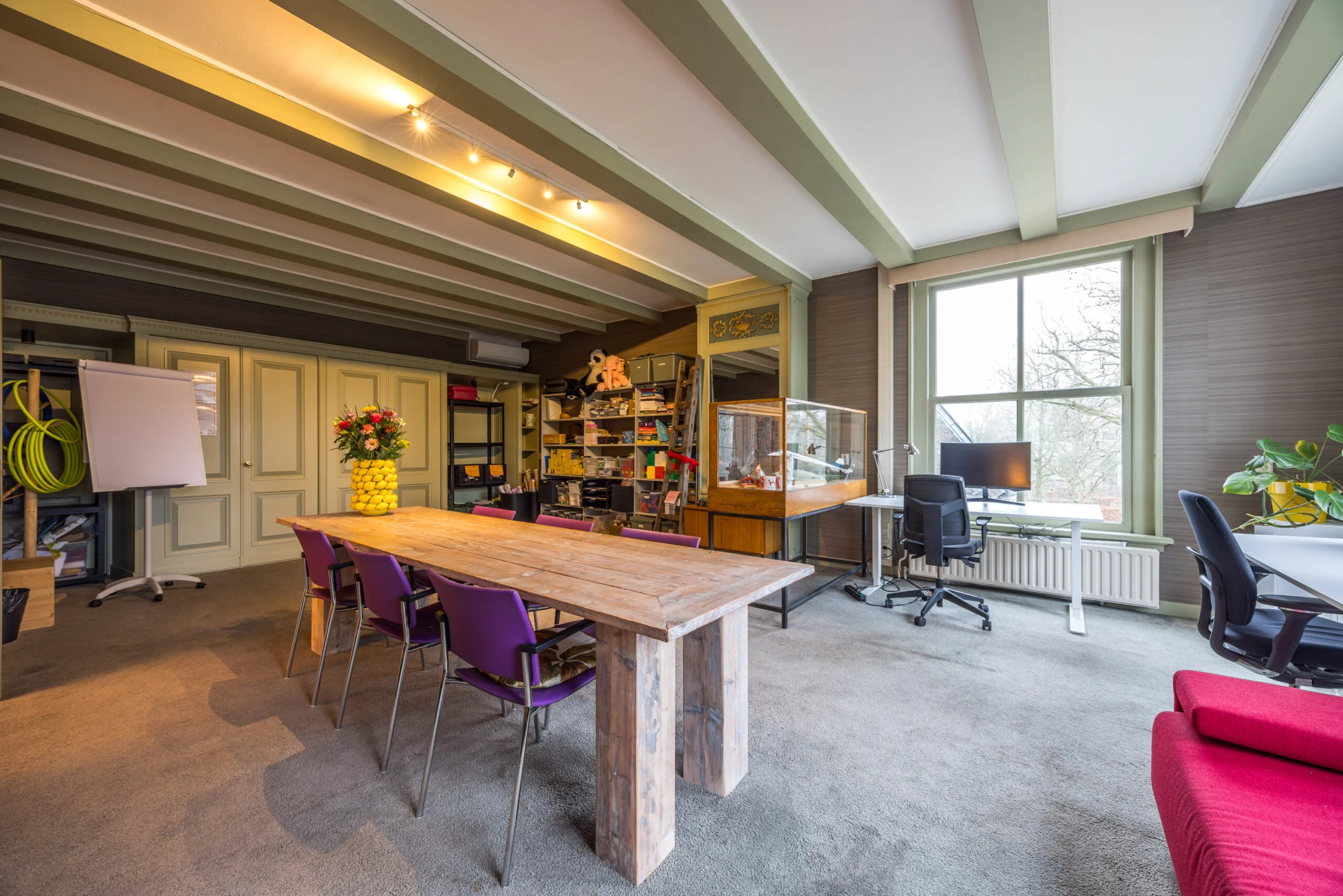 Ruim en kleurrijk ingericht kantoor met houten vergadertafel, paarse stoelen, bureau bij het raam en open opbergvakken.