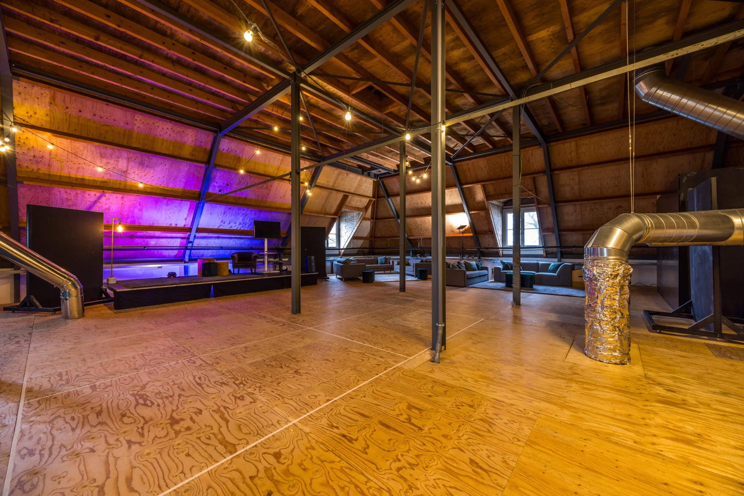 Ruime zolderverdieping met houten vloer, stalen constructie, een podium met paarse verlichting en loungehoek met banken.