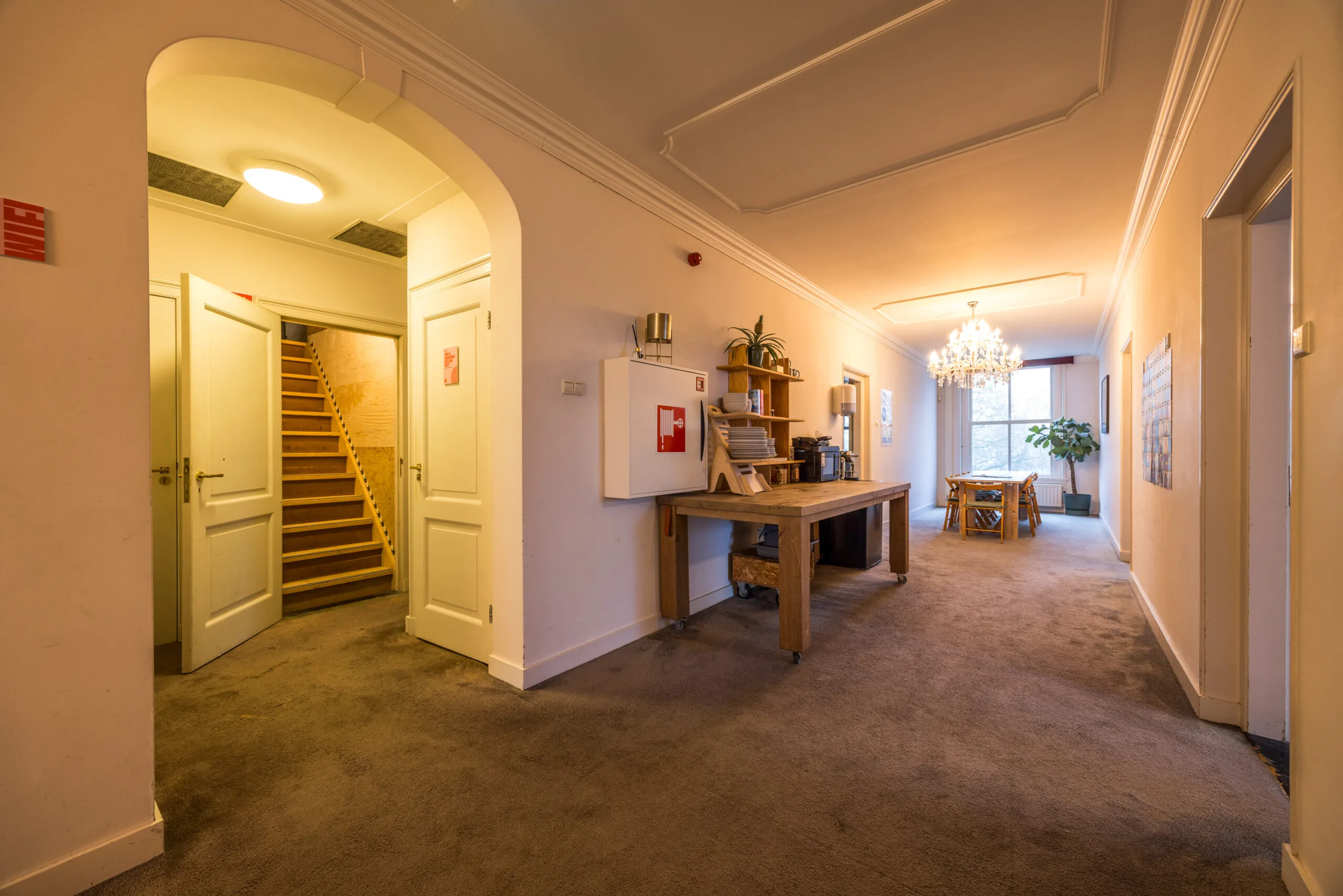 Brede gang in een huiselijke omgeving met een trapopgang, houten tafel met servies en een eetgedeelte aan het einde bij een raam.
