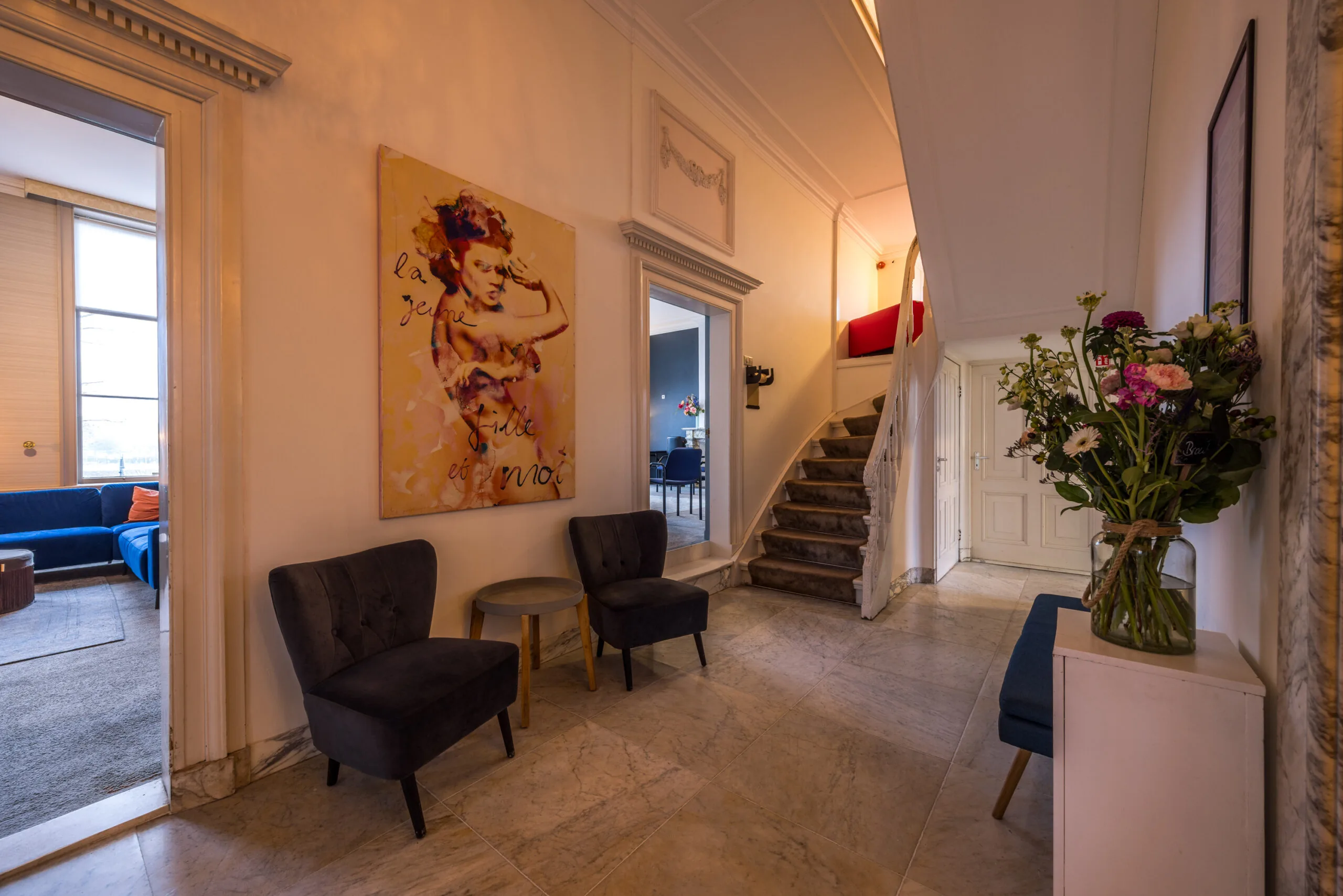 Entree van een elegant huis met marmeren vloer, kunst aan de muur, een trap en bloemen op een kastje.