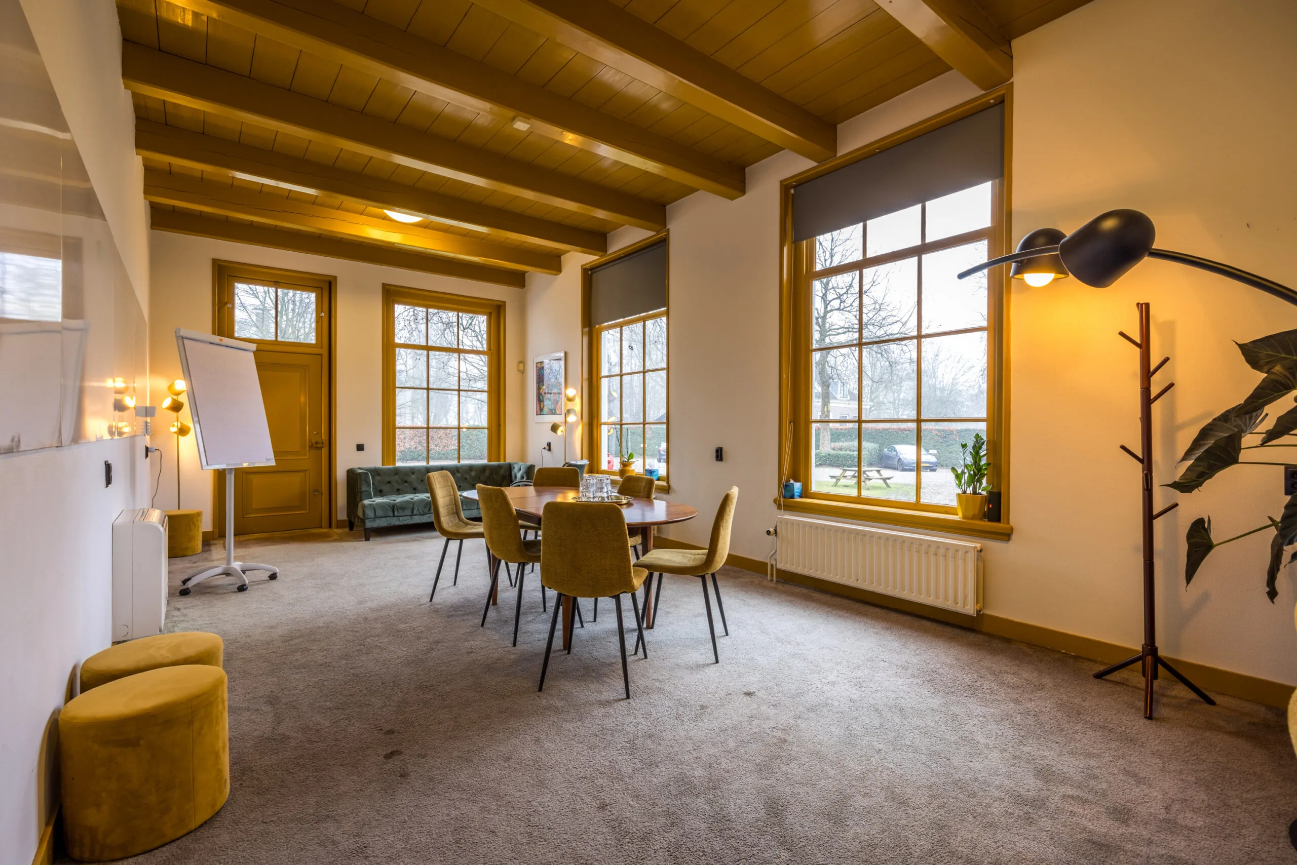 Vergaderruimte met een ronde tafel, zes gele stoelen, grote ramen en een flip-over in een sfeervolle, huiselijke setting.