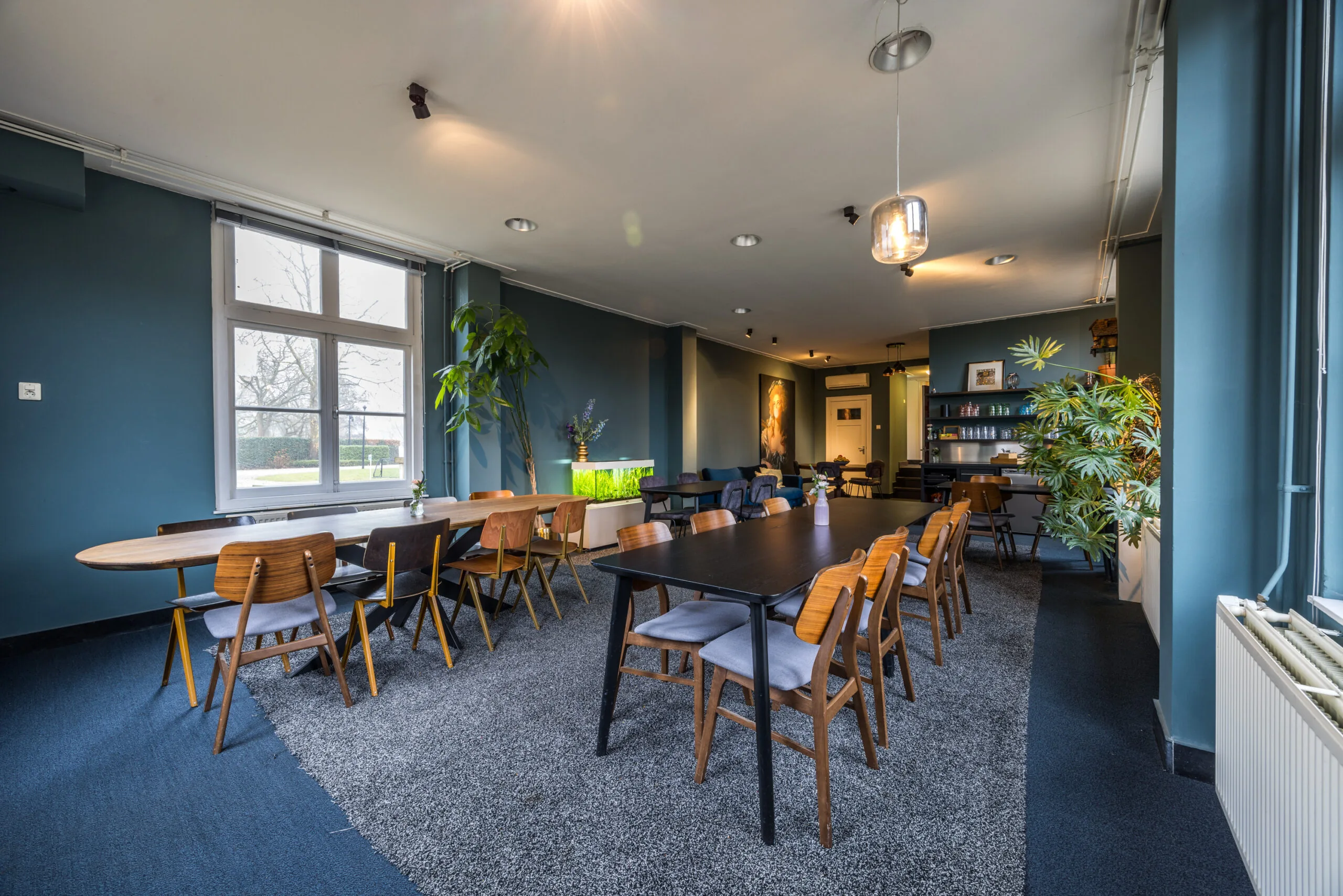 Modern ingerichte vergaderruimte met lange tafels, houten stoelen, groene planten en grote ramen met uitzicht op buiten.