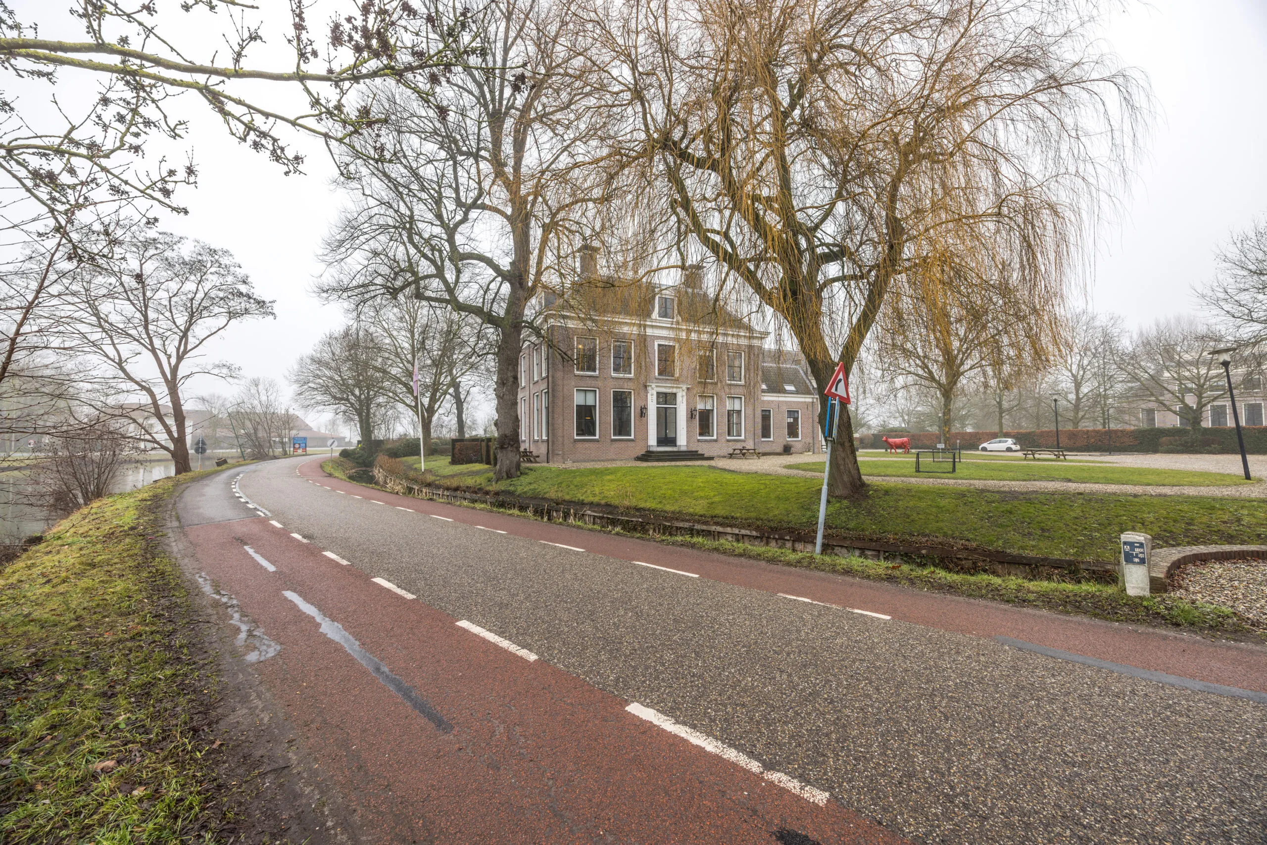 Een bochtige weg langs een gracht met een groot, klassiek herenhuis en kale bomen in een mistig landschap aan het Zandpad.