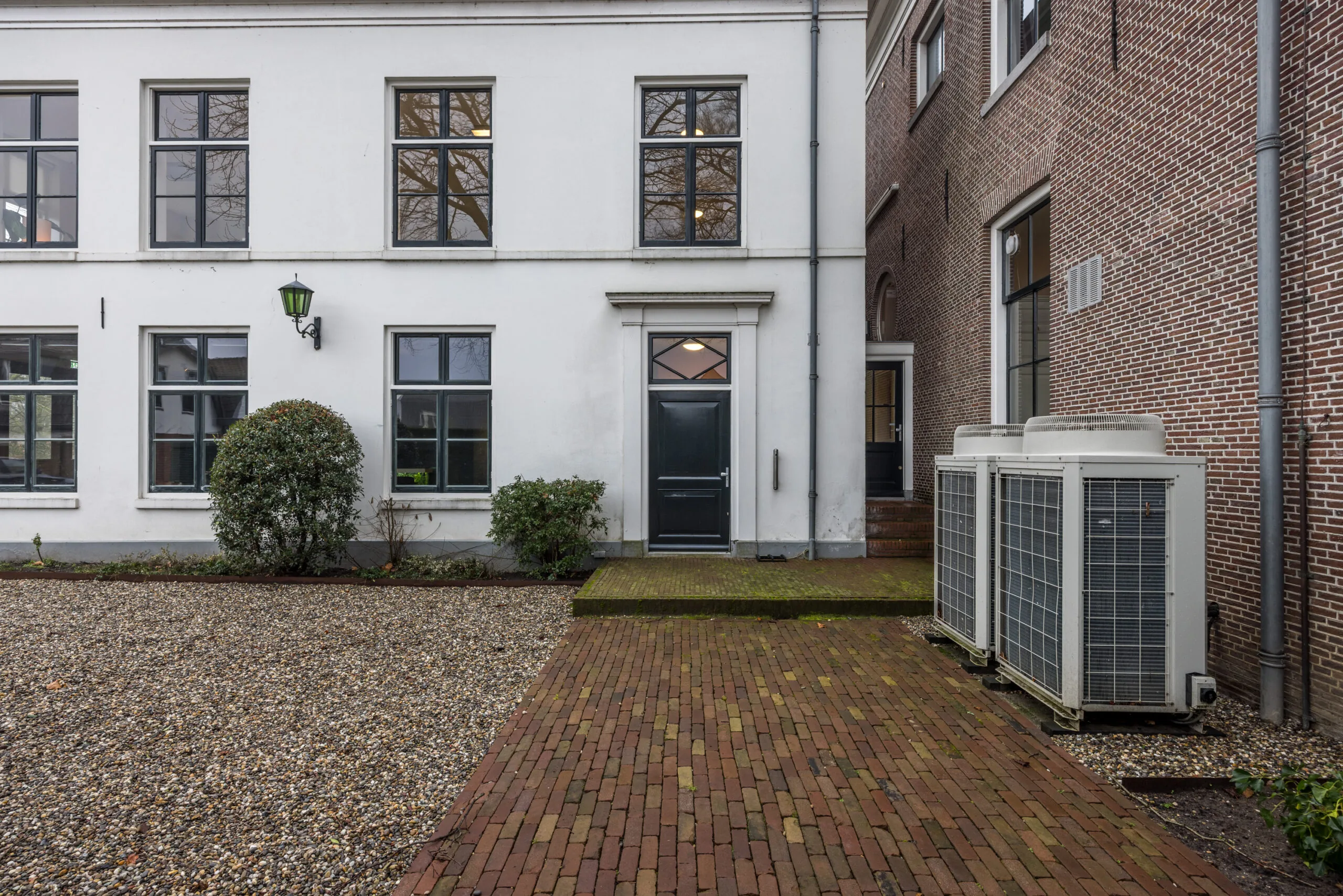 Binnenplaats met een witte gevel, zwarte deur, grindpad en airco-units aan de rechterkant.