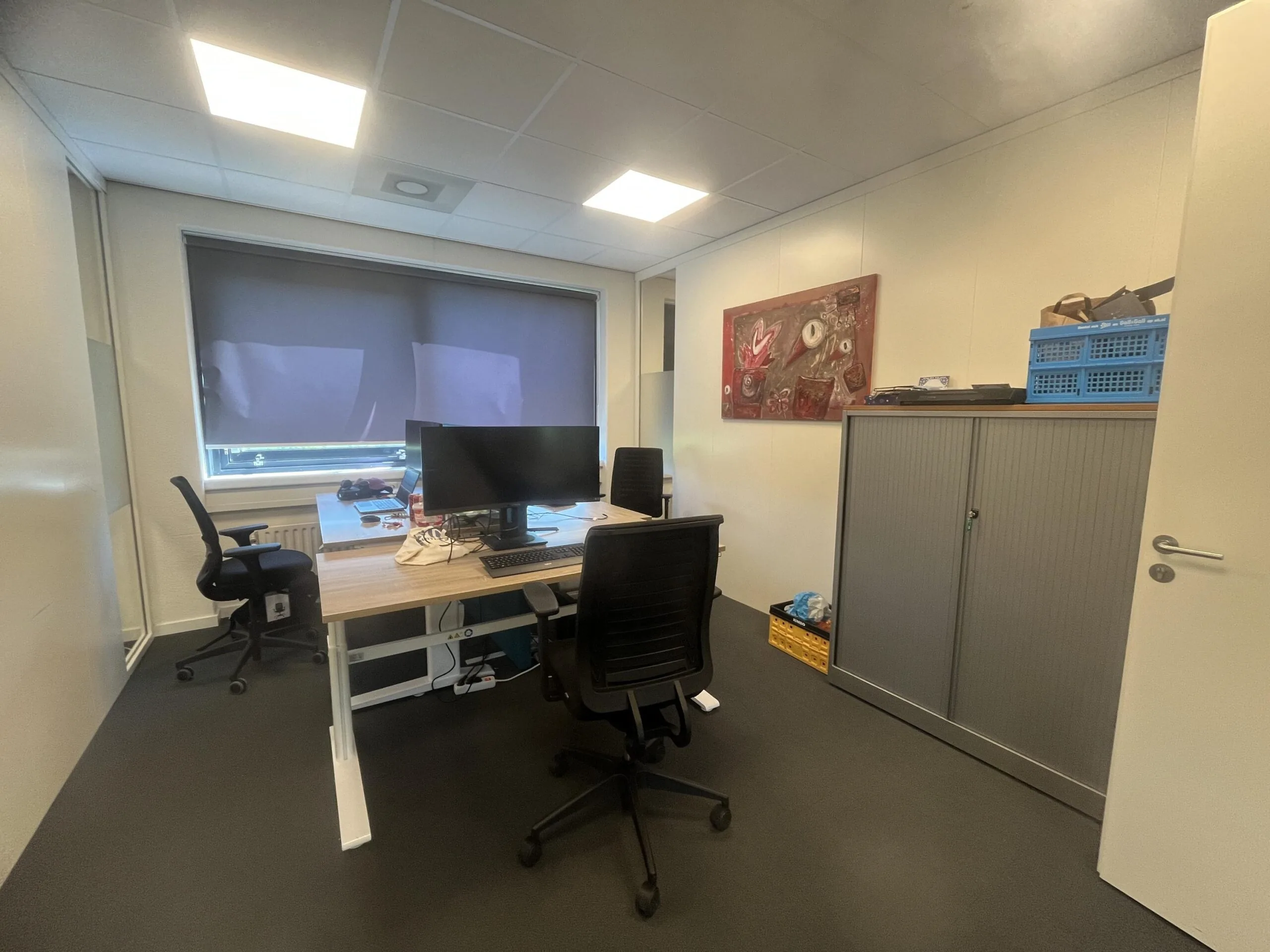Klein kantoor met twee bureaus, computerschermen, een gesloten rolgordijn en een kast met blauwe opbergkratten.