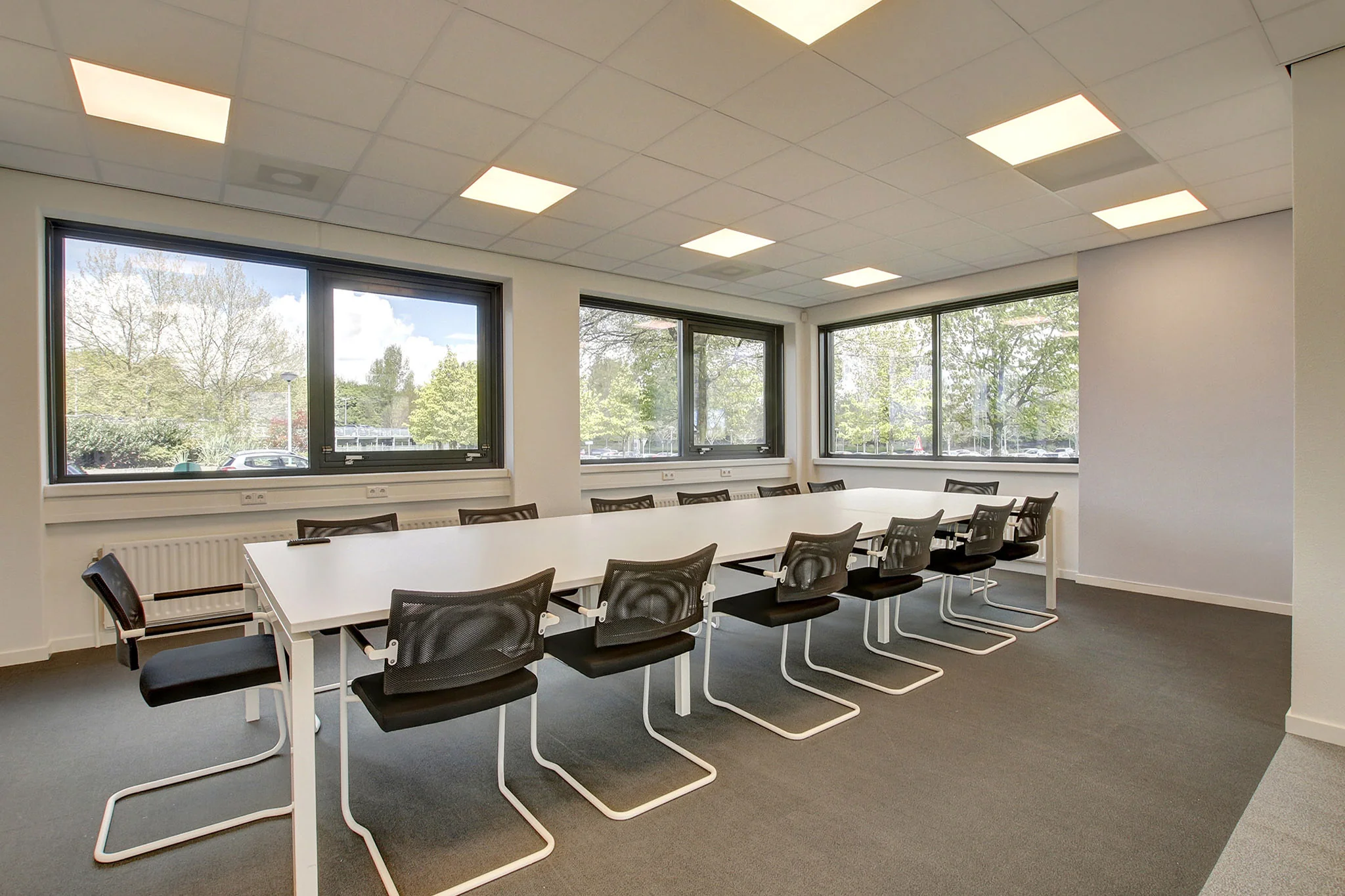 Vergaderruimte met een lange witte tafel, zwarte stoelen en grote ramen aan twee zijden in een kantoorpand aan de Nevelgaarde.