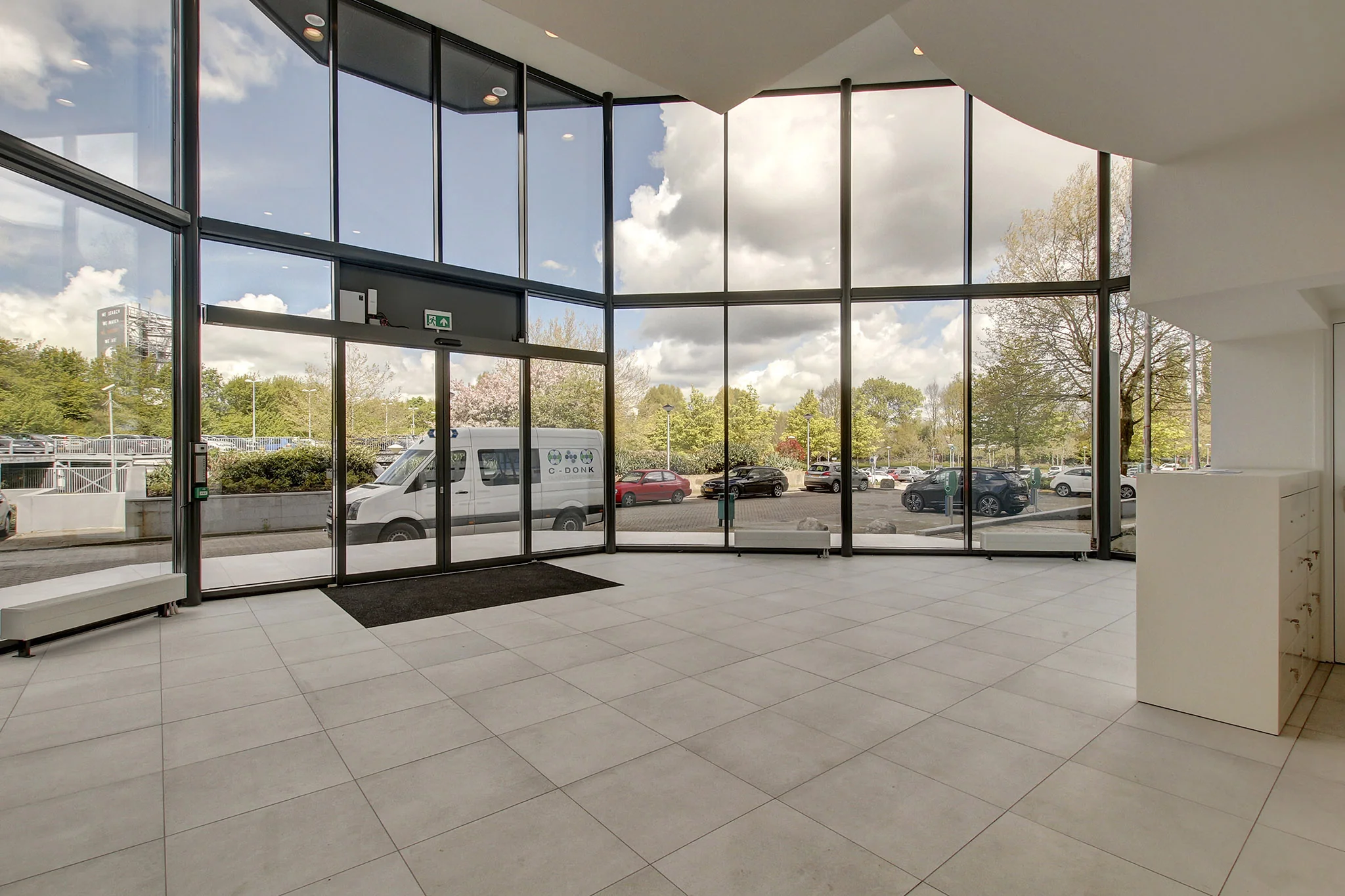 Ruime, lichte entree met hoge glazen pui en uitzicht op parkeerplaats aan de Nevelgaarde.