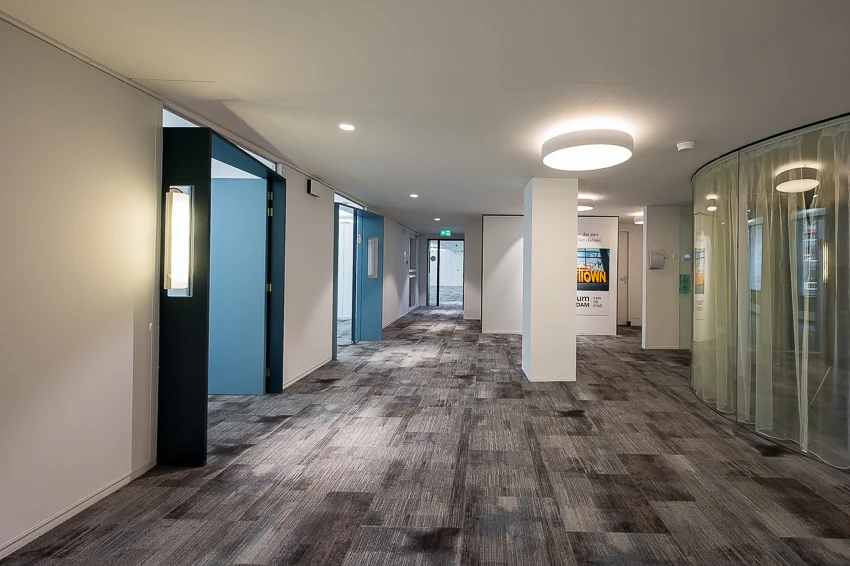 Een moderne gang in een kantoorgebouw met blauwe deuren, plafondverlichting en gebogen glazen wand.