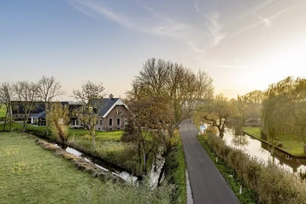 Landelijke weg langs het water in Gein-Zuid met een boerderij en herfstige bomen bij zonsondergang.