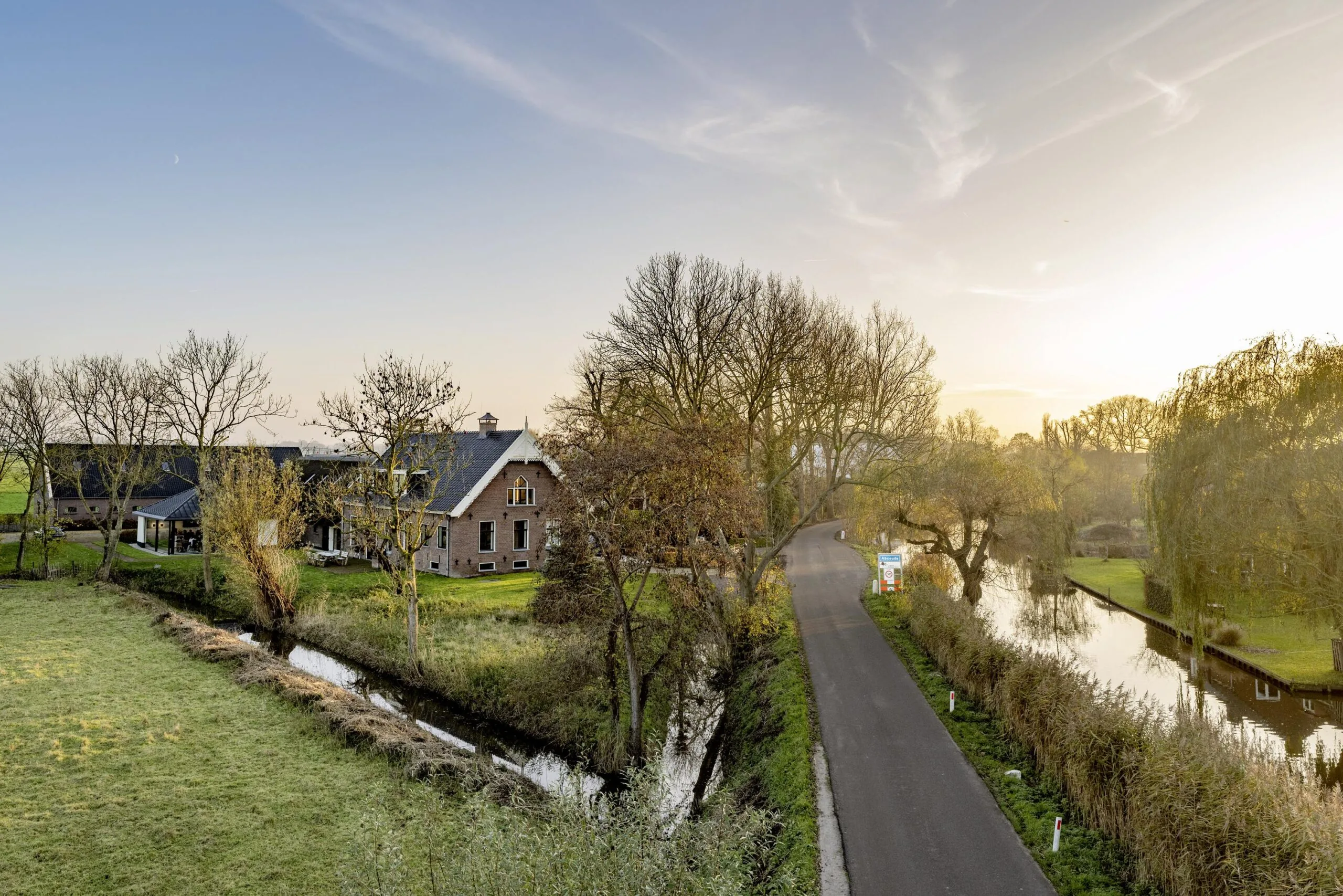 Landelijke weg langs het water in Gein-Zuid met een boerderij en herfstige bomen bij zonsondergang.
