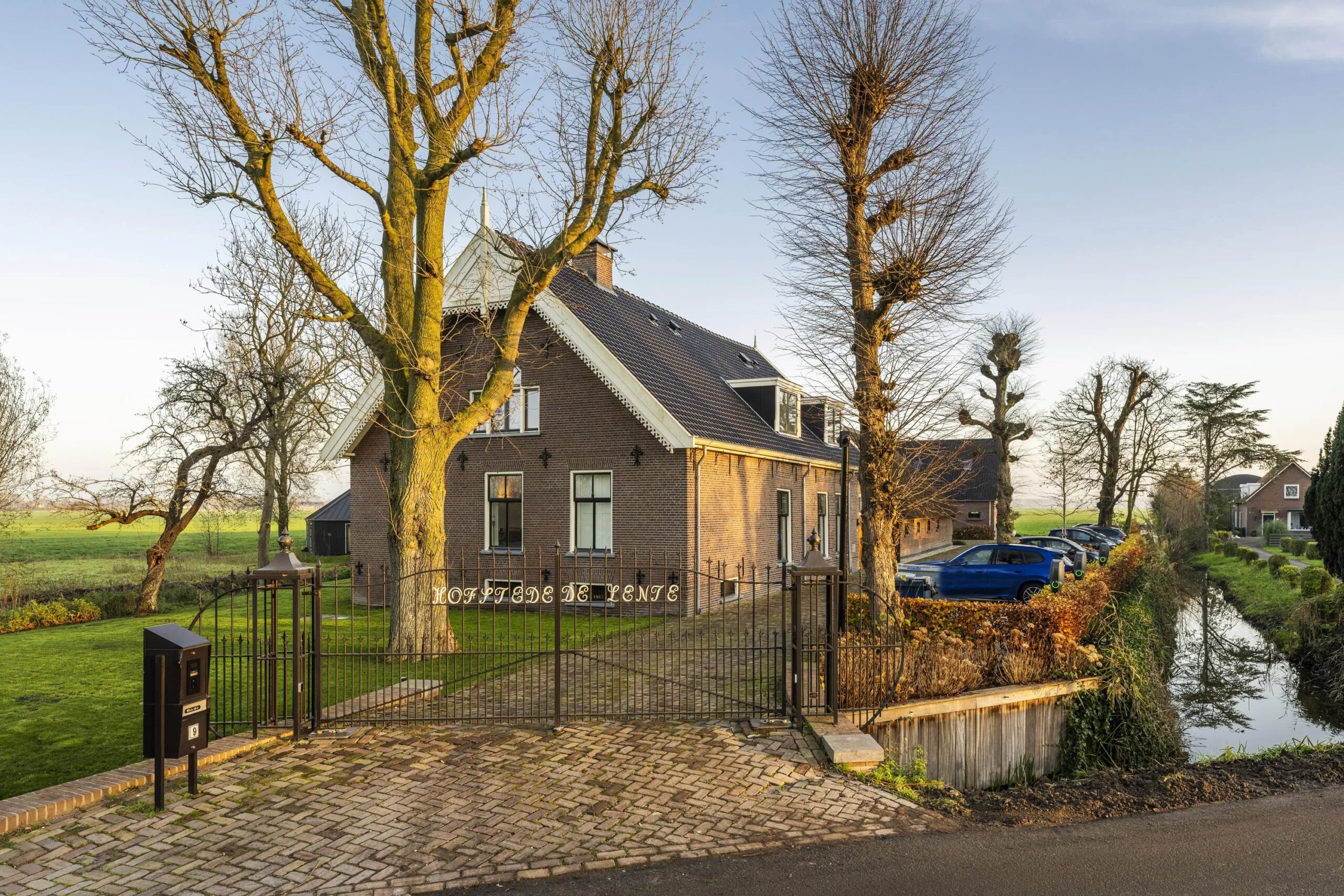 Grote landelijke woning genaamd "Hofstede de Vente" met oprijlaan, geparkeerde auto's en uitzicht op het omliggende polderlandschap.
