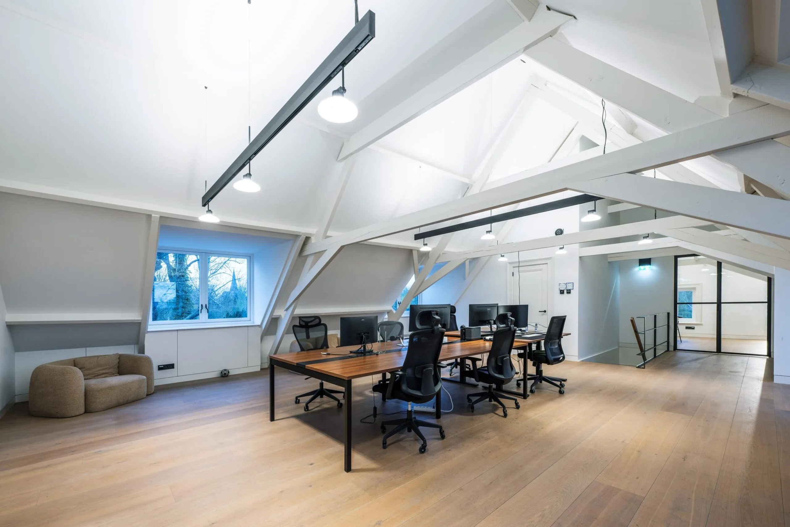 Modern kantoor op zolderverdieping met houten vloer, schuine witte balken en acht werkplekken aan een grote houten tafel.