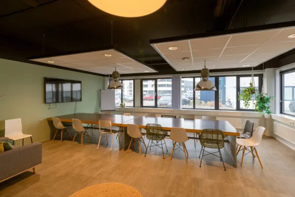 Vergaderruimte aan de Kobaltweg met een lange tafel, diverse stoelen, een flatscreen, en grote ramen met uitzicht op een parkeerterrein.