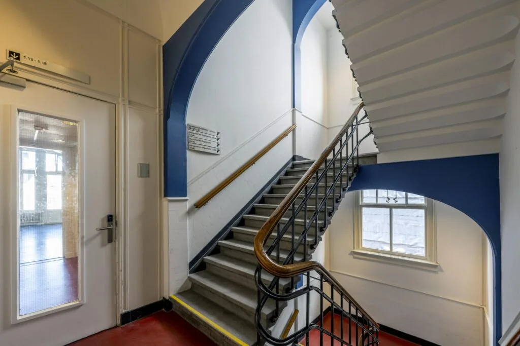 Trap in een gebouw met blauwe accenten en een bord met route-indicaties aan de muur.