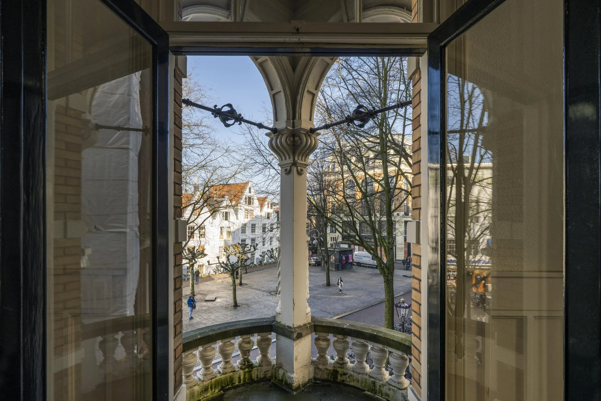 Uitzicht vanuit een open raam op een straat met traditionele gebouwen en kale bomen in de Handboogstraat.