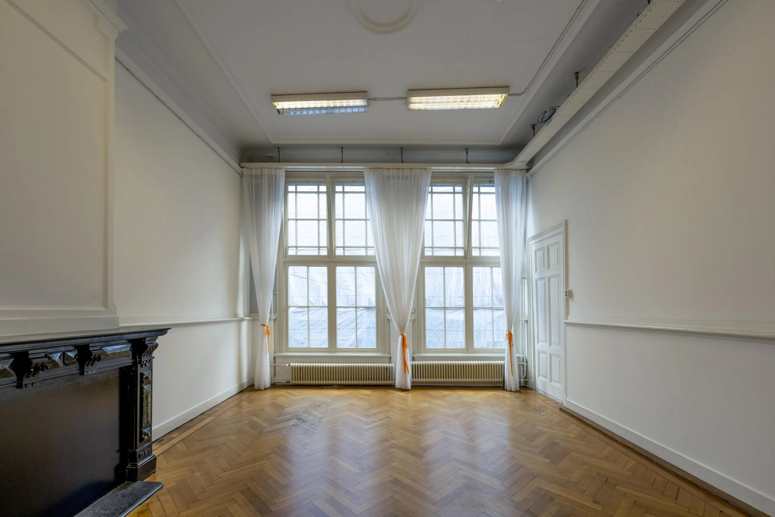Een lege kamer met houten vloer, grote ramen en een zwarte schoorsteenmantel in de Handboogstraat.