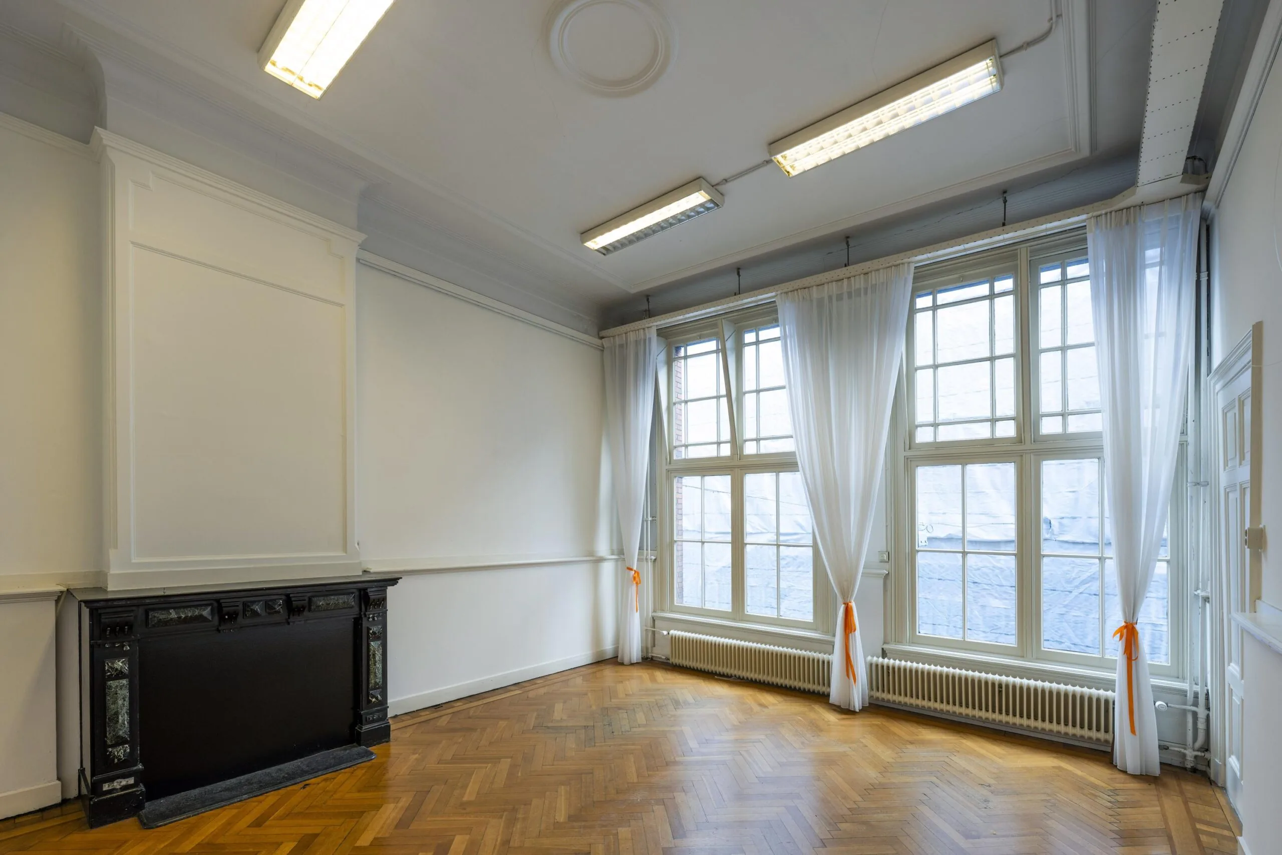 Een lege kamer met grote ramen, witte gordijnen, houten vloer en een zwarte schouw.