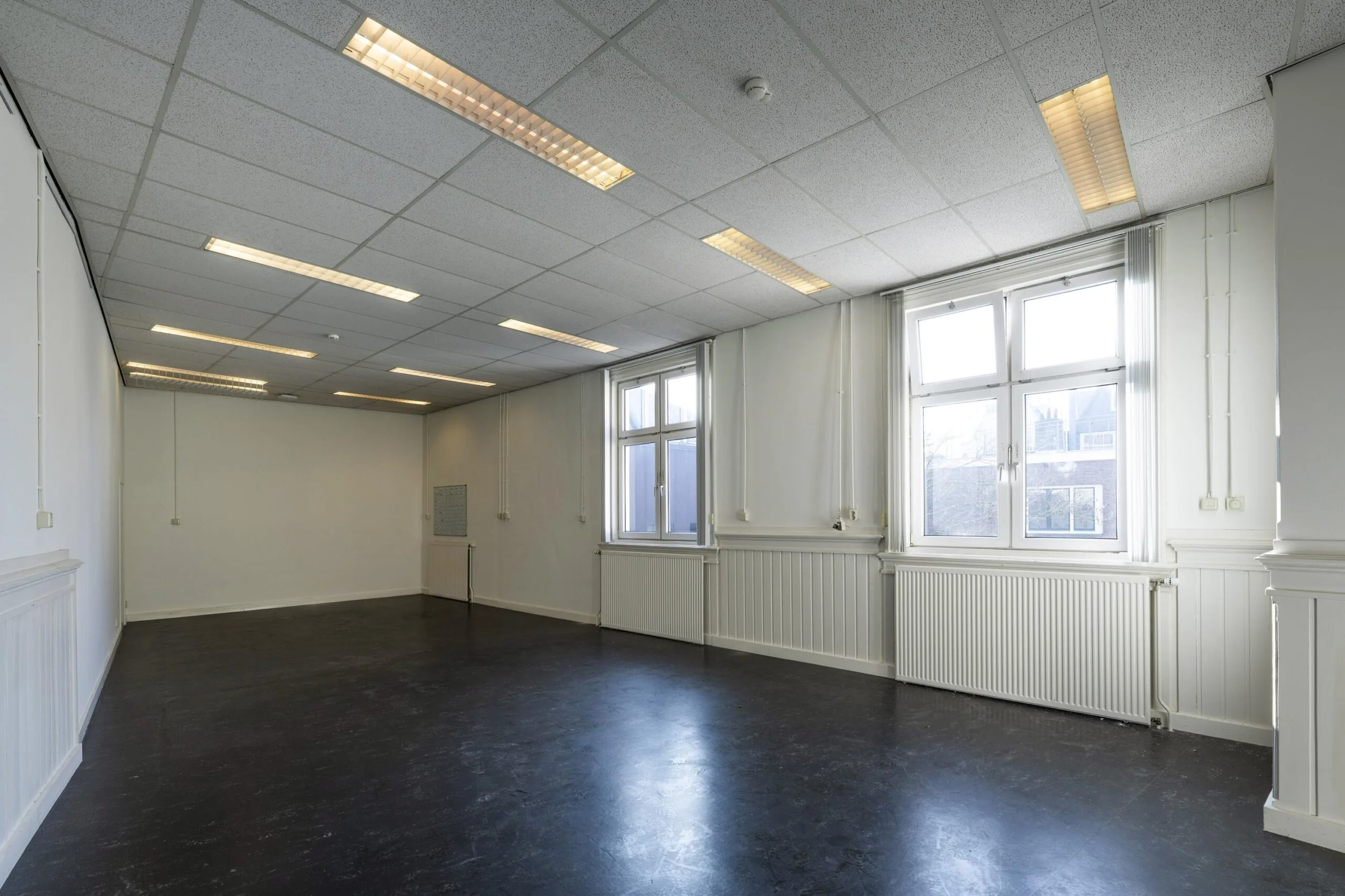 Leeg kantoor met witte muren, twee grote ramen en TL-verlichting aan het plafond.