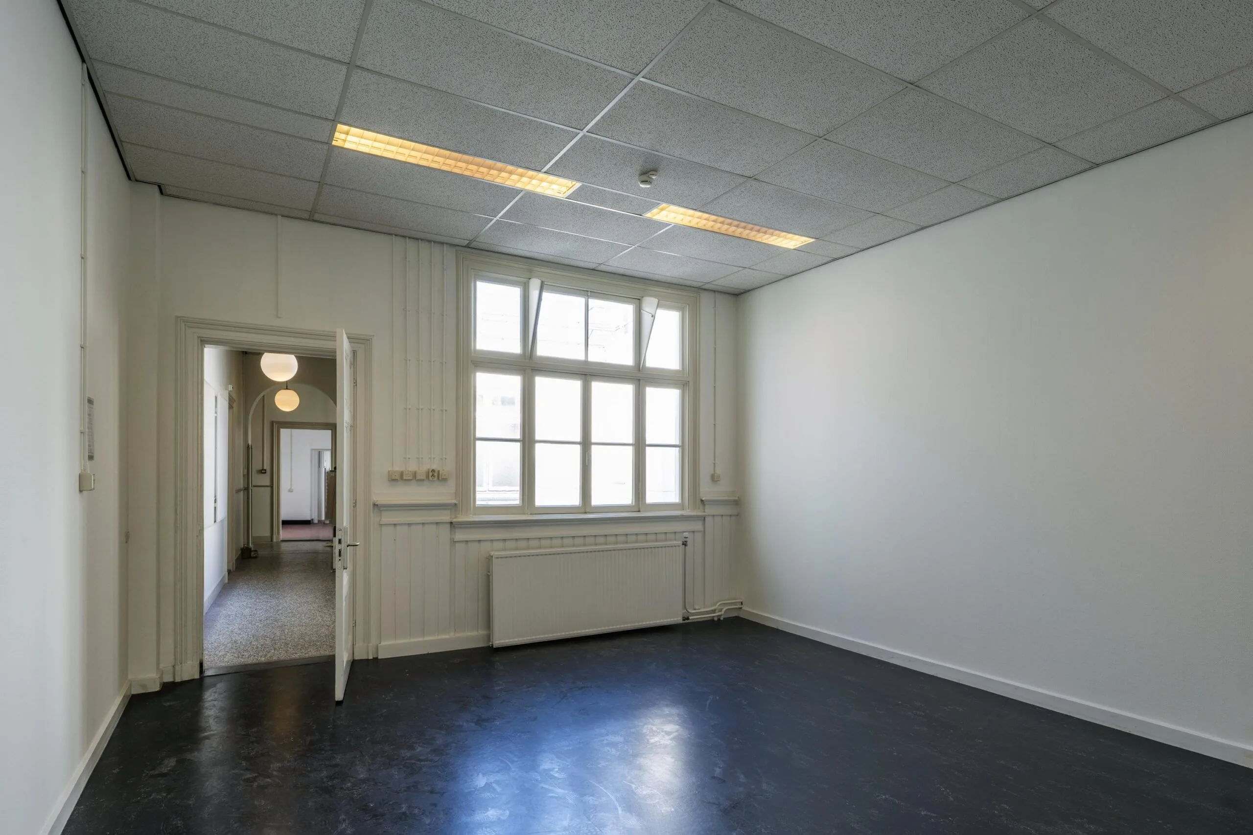 Een lege kamer met een groot raam en een open deur naar een gang.