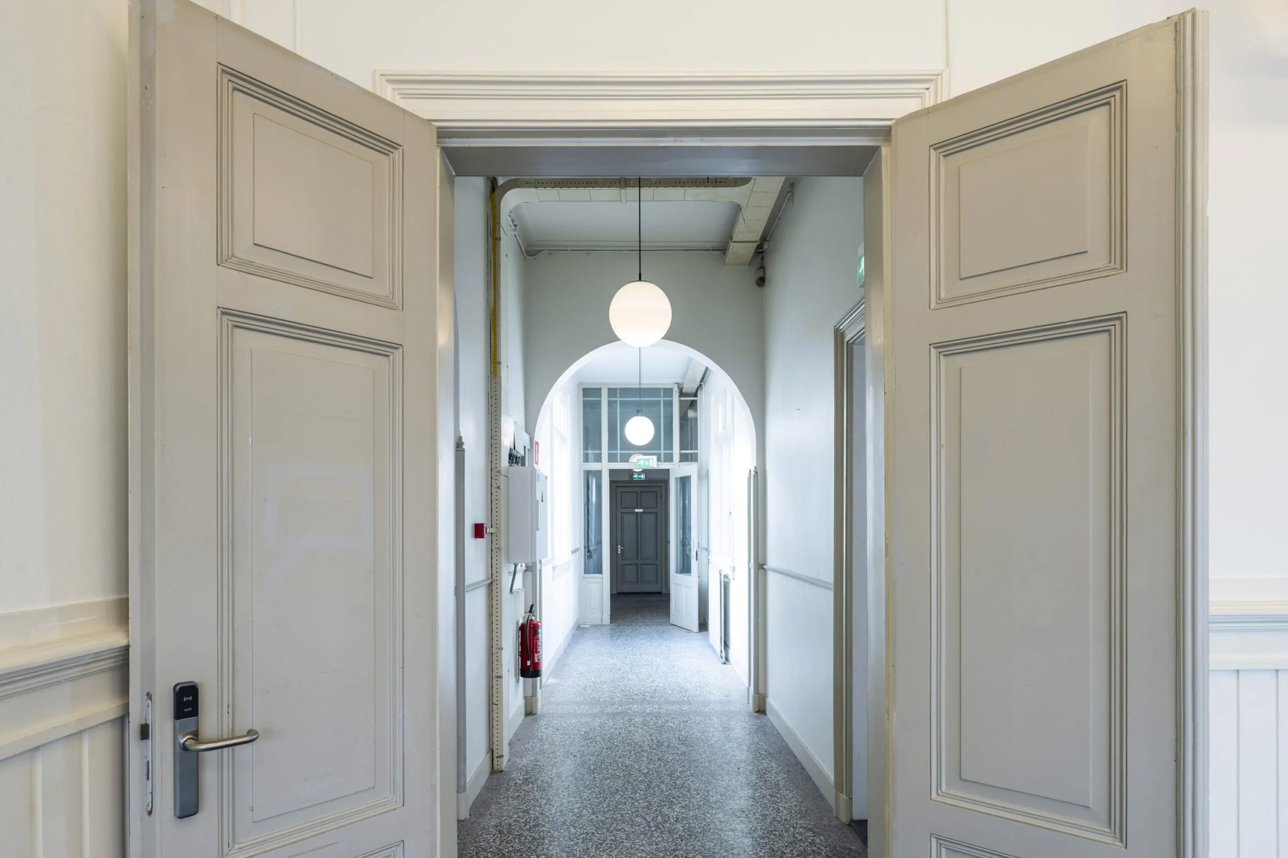 Een open deur leidt naar een lange gang met een bolvormige lamp aan het plafond.