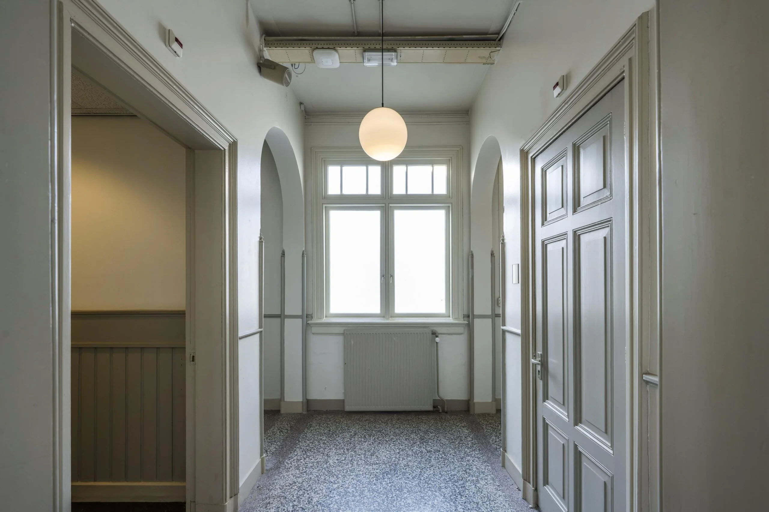 Een lichte gang met een groot raam, een ronde hanglamp, en grijze houten deuren in de Handboogstraat.