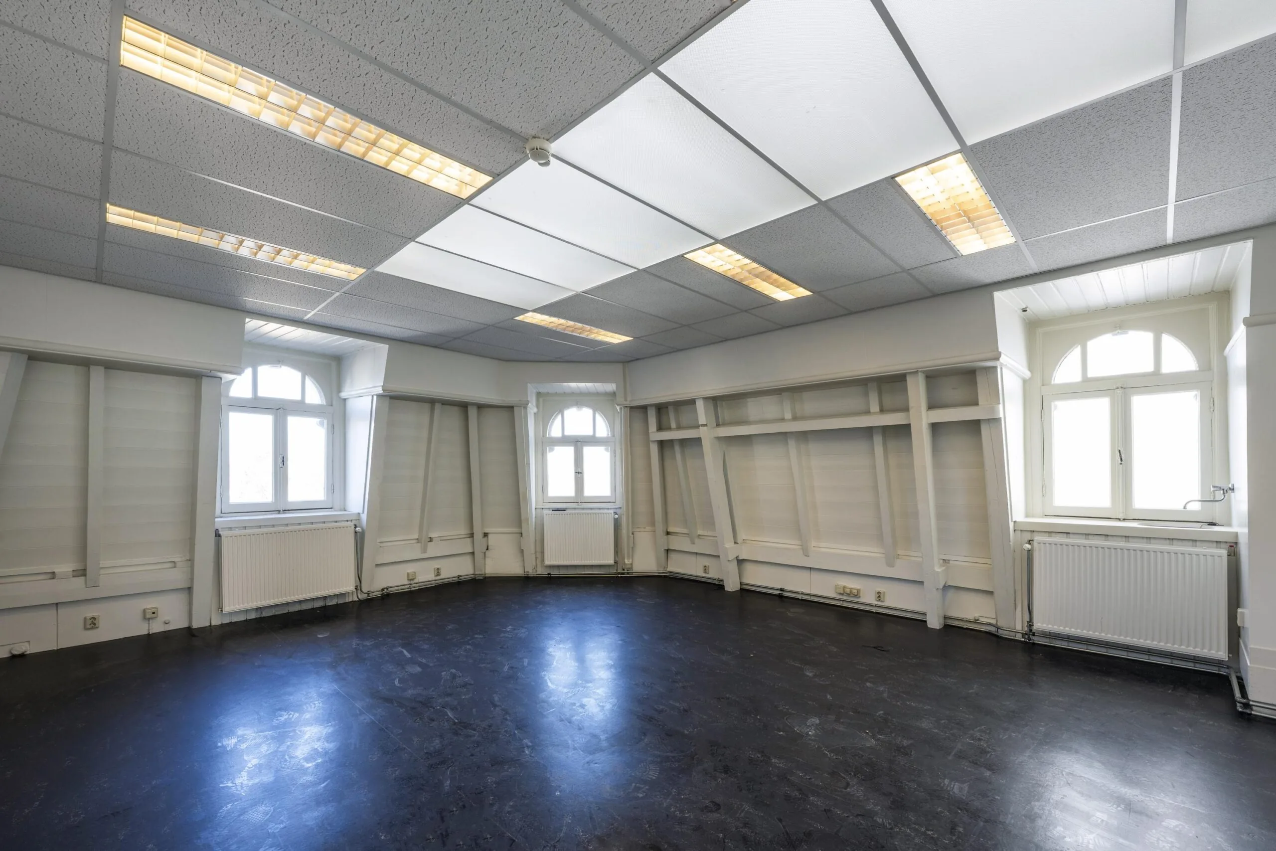Een lege kamer met drie grote ramen, witte muren en een systeemplafond met tl-verlichting.