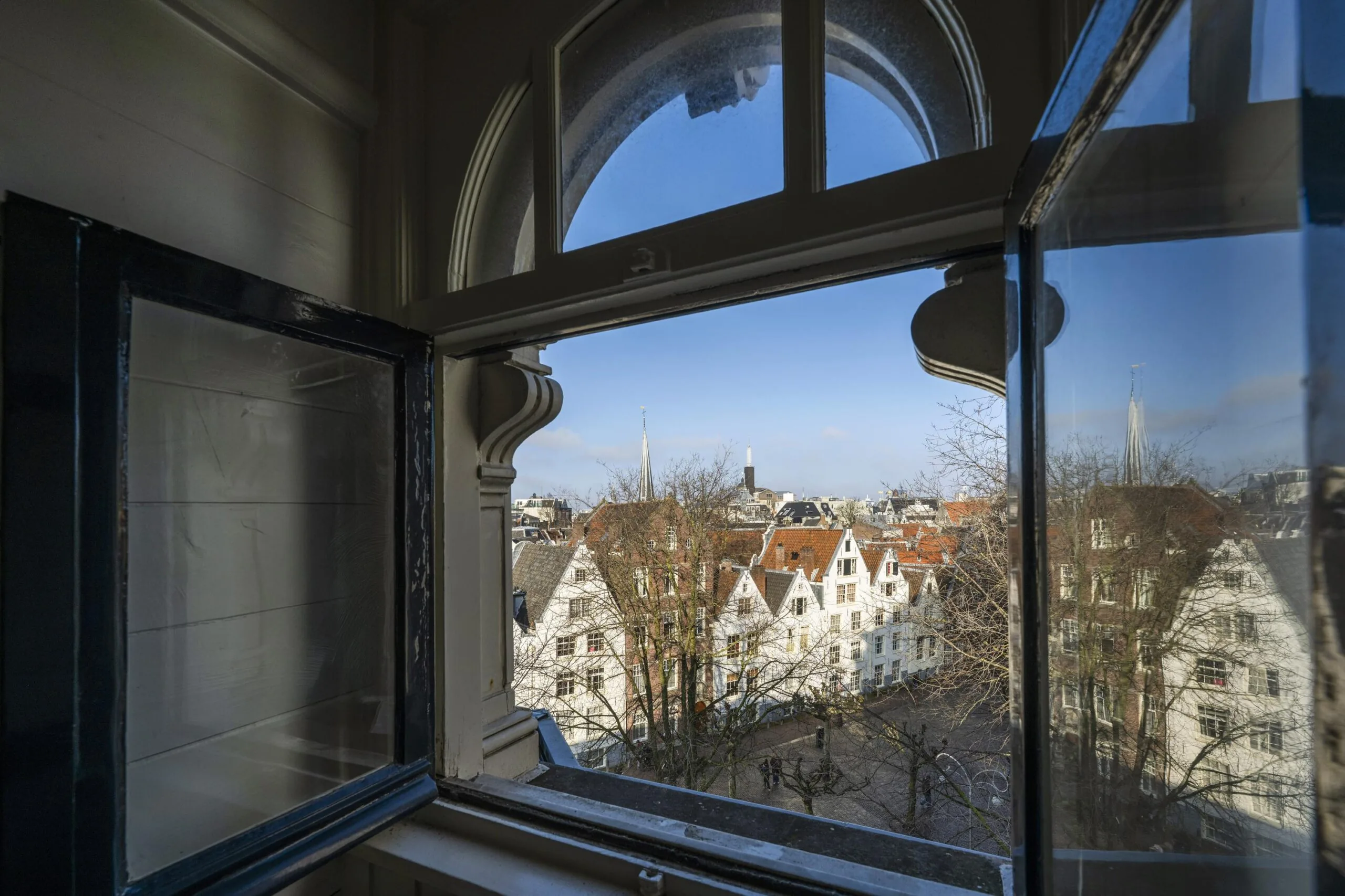 Uitzicht vanuit een open raam op historische gebouwen aan de Handboogstraat met een blauwe lucht in de achtergrond.