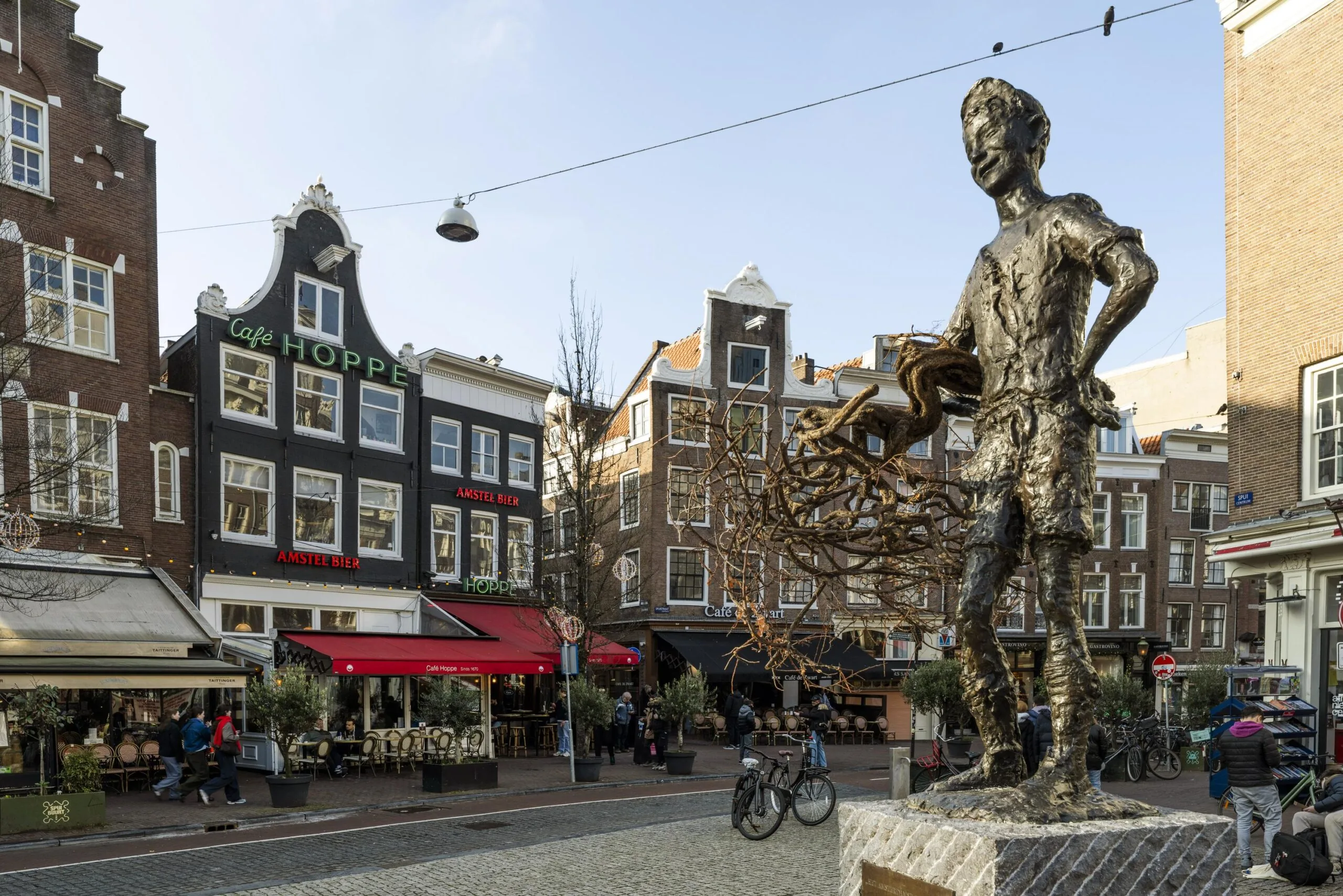 Standbeeld van een persoon op een levendige straat met cafés en historische gebouwen op de achtergrond.