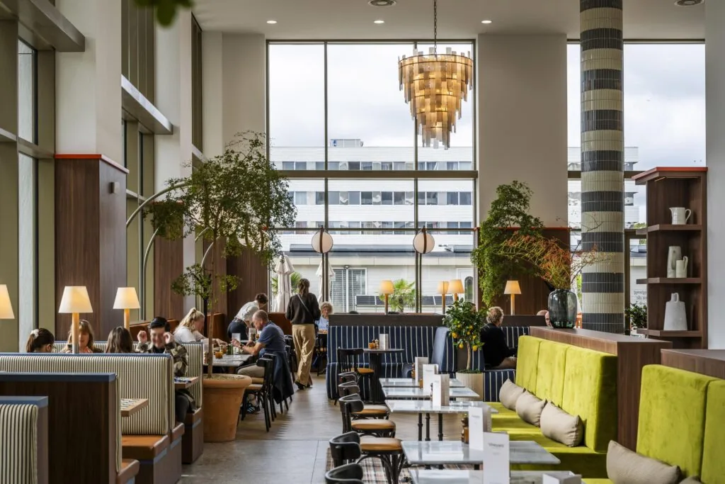 Een modern ingericht restaurant met grote ramen, groene planten en mensen die aan tafels zitten.