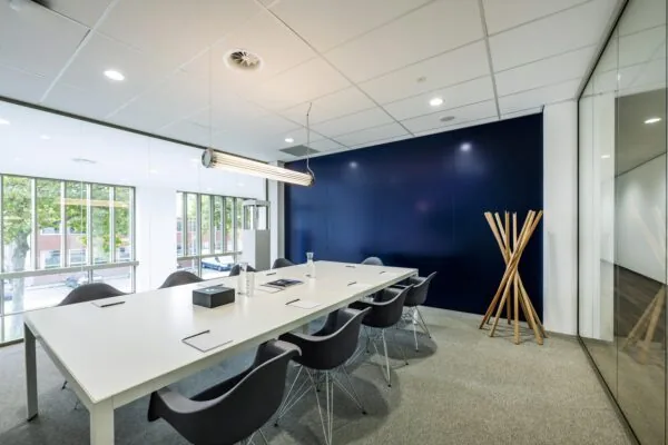 Een moderne vergaderruimte met een grote witte tafel en grijze stoelen, omgeven door glazen wanden en een blauwe muur.