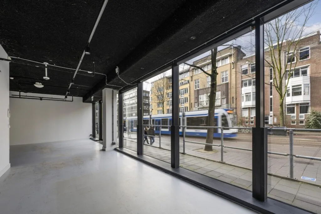 Een lege ruimte met grote ramen aan de Roetersstraat, met zicht op een rijdende tram en omringende gebouwen.