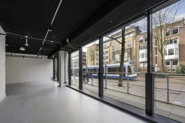Een lege ruimte met grote ramen aan de Roetersstraat, met zicht op een rijdende tram en omringende gebouwen.