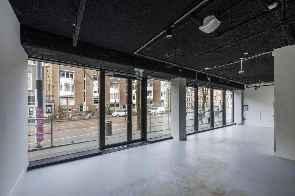 Een lege ruimte met grote ramen aan de Roetersstraat, uitkijkend op gebouwen en een straat met fietsen.