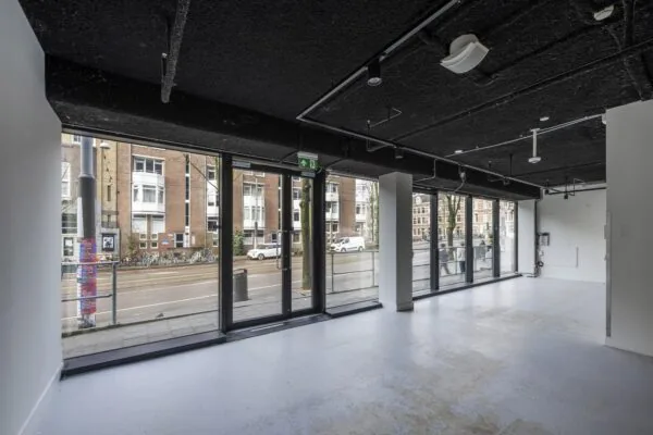 Een lege ruimte met grote ramen aan de Roetersstraat, uitkijkend op gebouwen en een straat met fietsen.