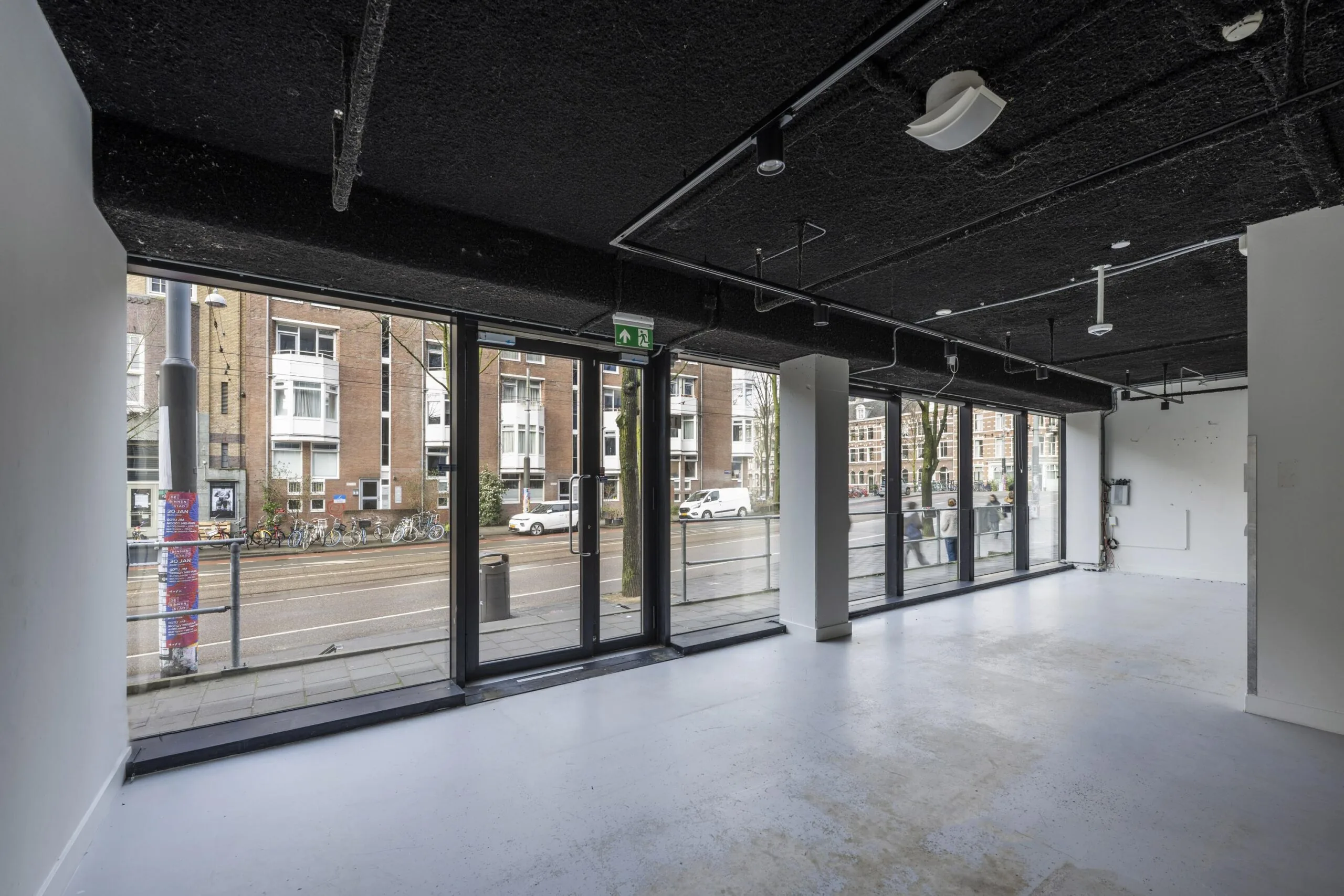 Een lege ruimte met grote ramen aan de Roetersstraat, uitkijkend op gebouwen en een straat met fietsen.