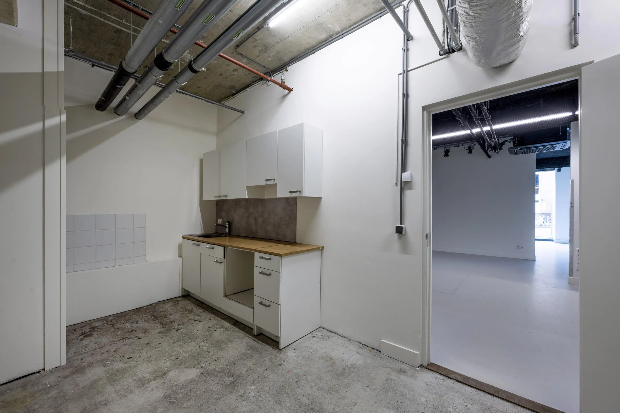 Een kleine keukenruimte met witte kasten en leidingen aan het plafond in een industrieel ogend gebouw.