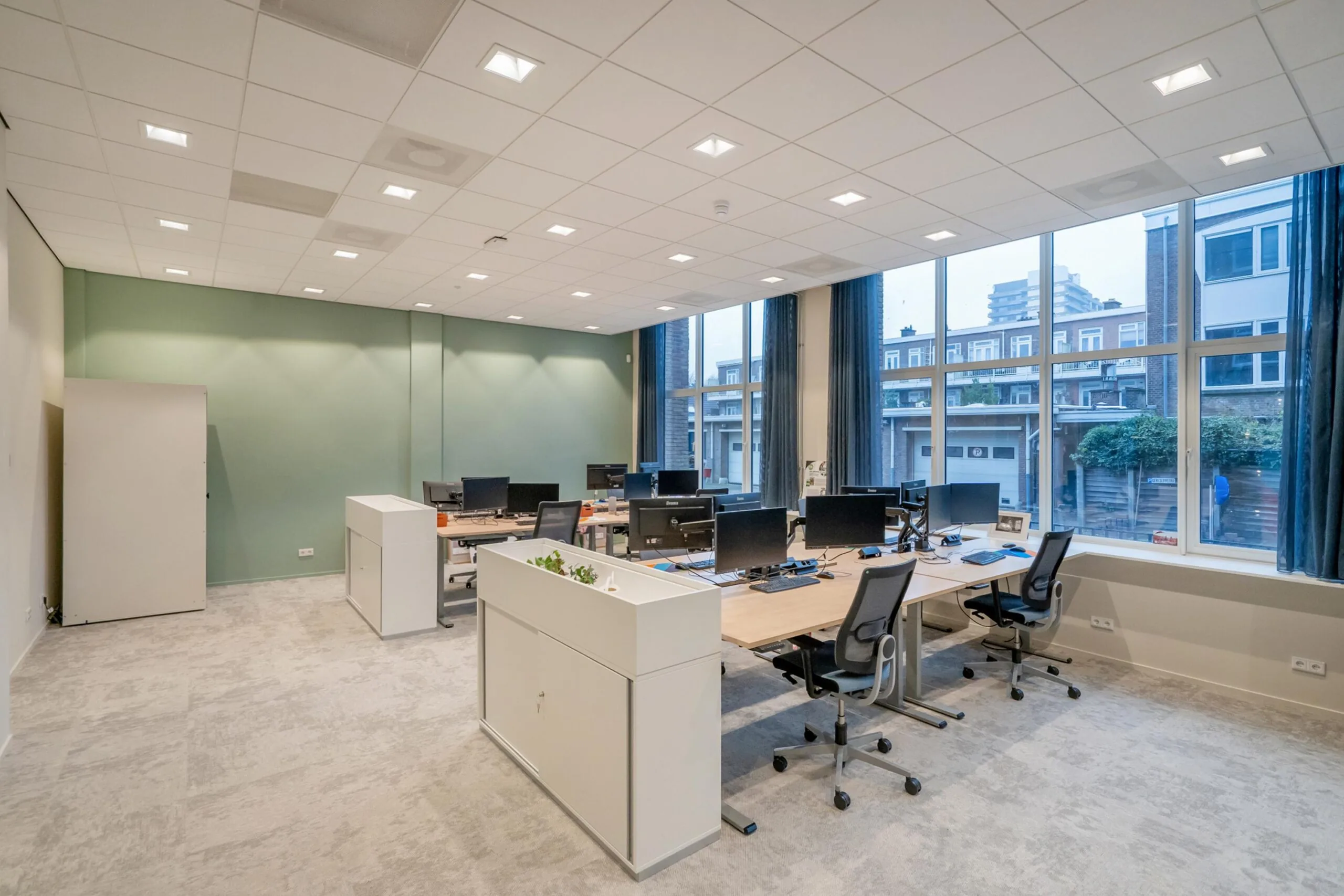 Een modern, licht kantoor met bureaus, bureaustoelen en computerschermen, met uitzicht op buiten.