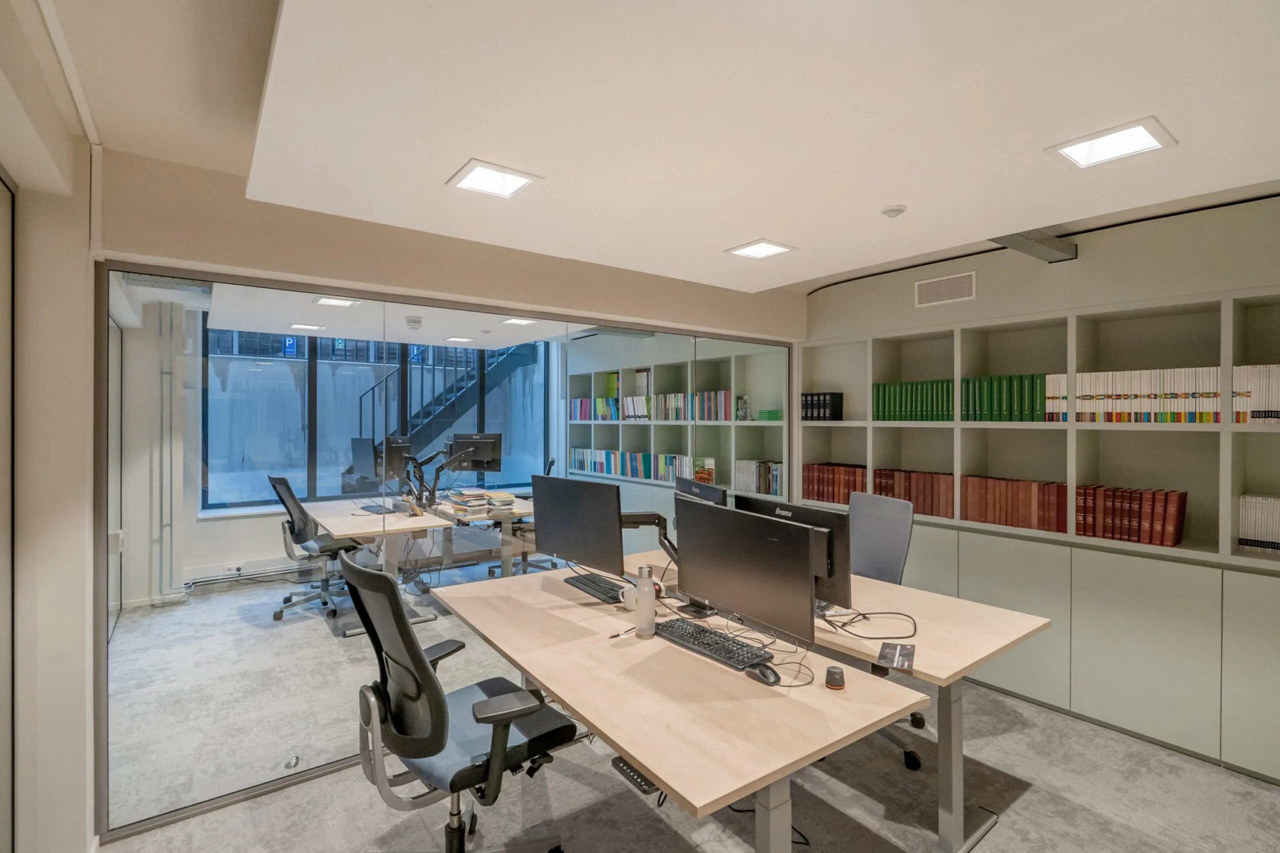 Een moderne kantoorruimte met bureaus, computers en een grote boekenplank aan de muur.