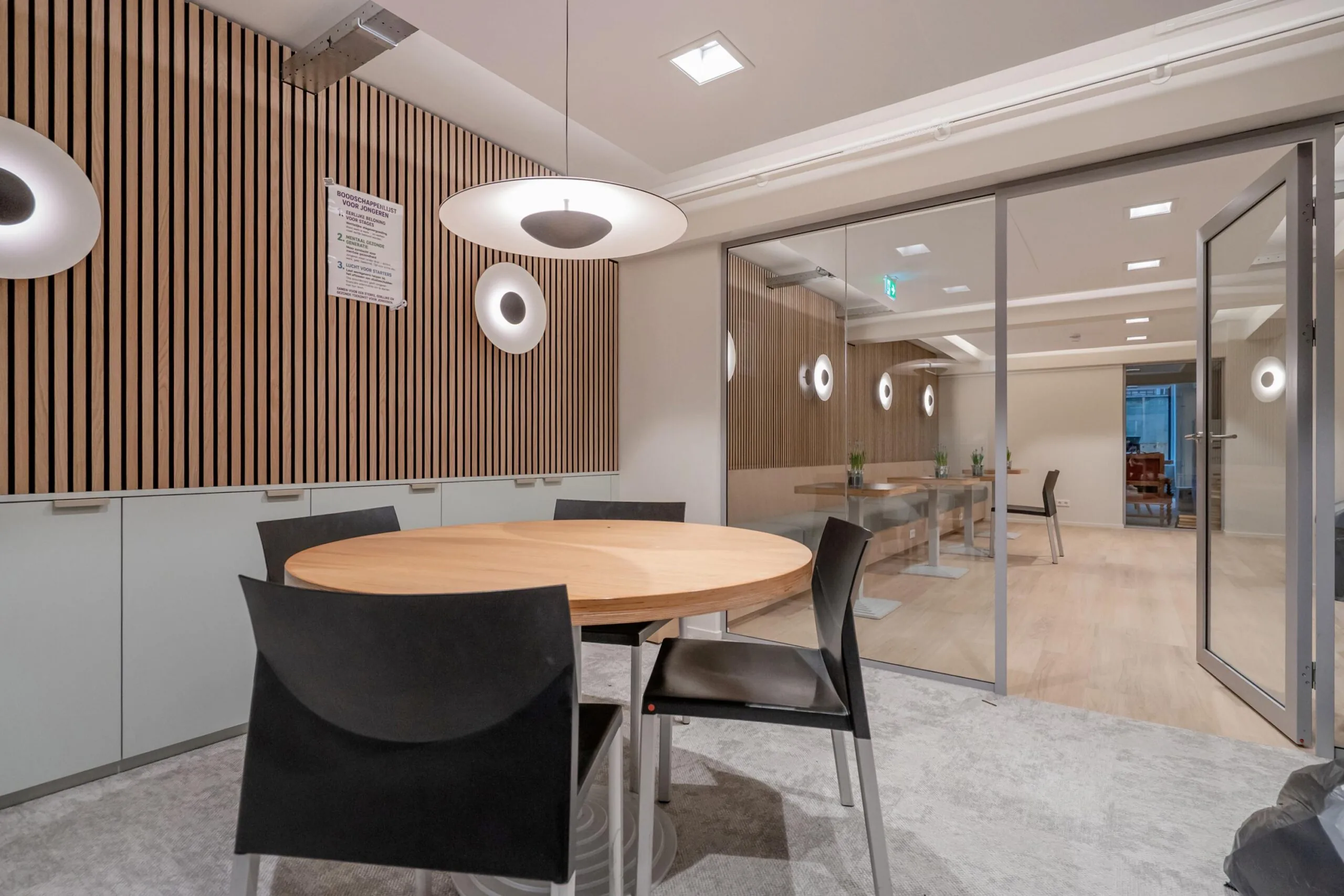 Een moderne vergaderruimte met een ronde houten tafel, zwarte stoelen en houten lambrisering op de muur.