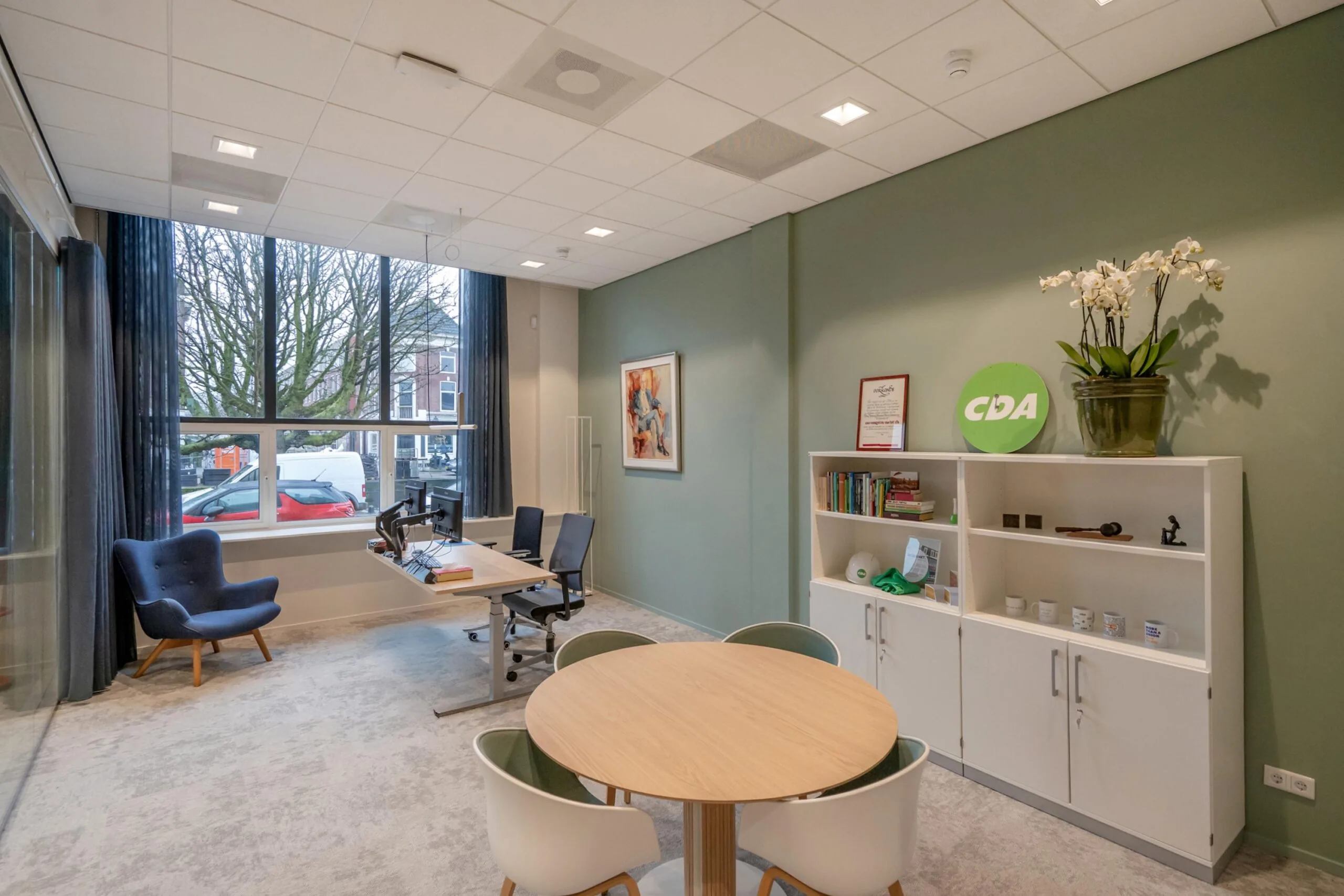 Een modern kantoor met een grote raam, een bureau, een zitgedeelte en een kast met een CDA-logo.