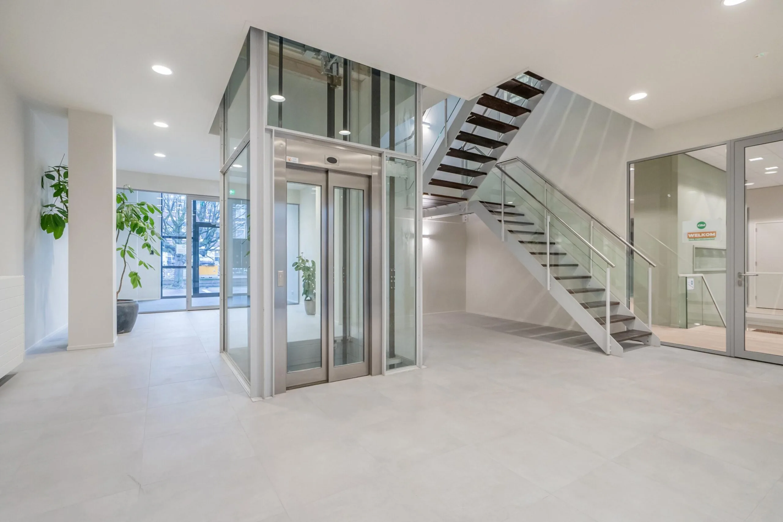 Een moderne, lichte hal met een glazen lift en een sierlijke trap.