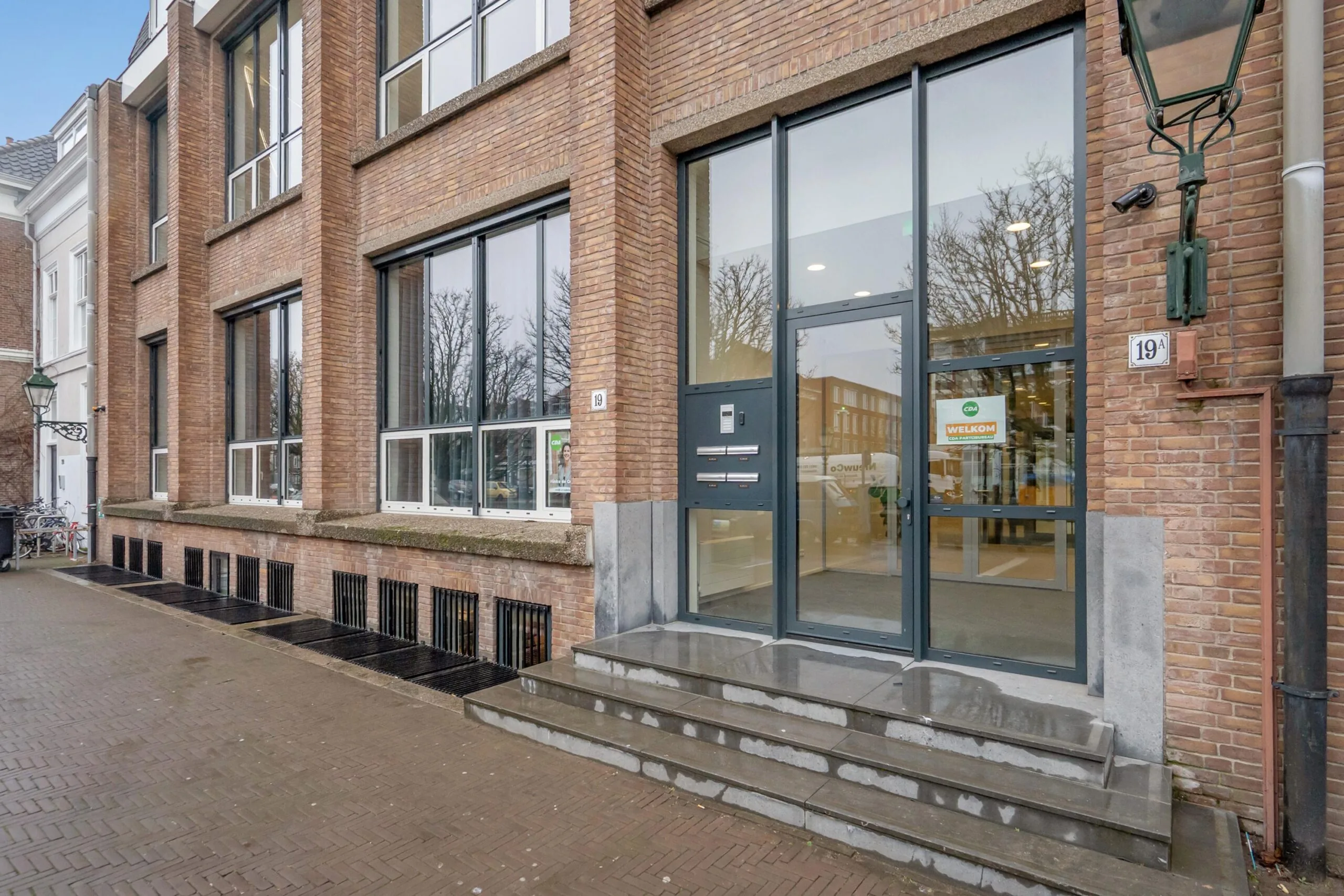 De afbeelding toont een bakstenen gebouw met een glazen entree en een bordje "Welkom" naast huisnummer 19.