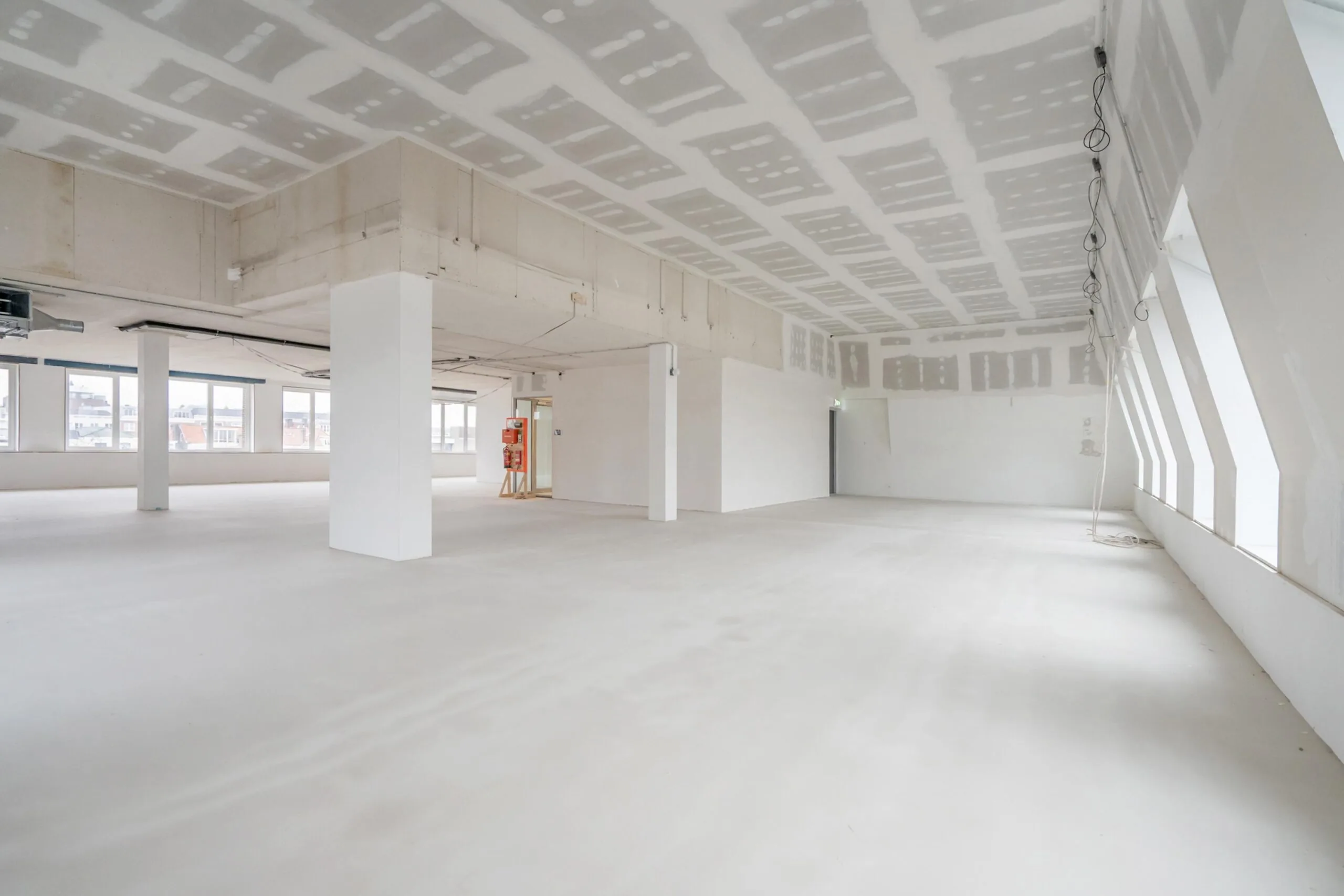 Lege ruimte in een onafgewerkt kantoor met grote ramen en zichtbare plafondconstructie.