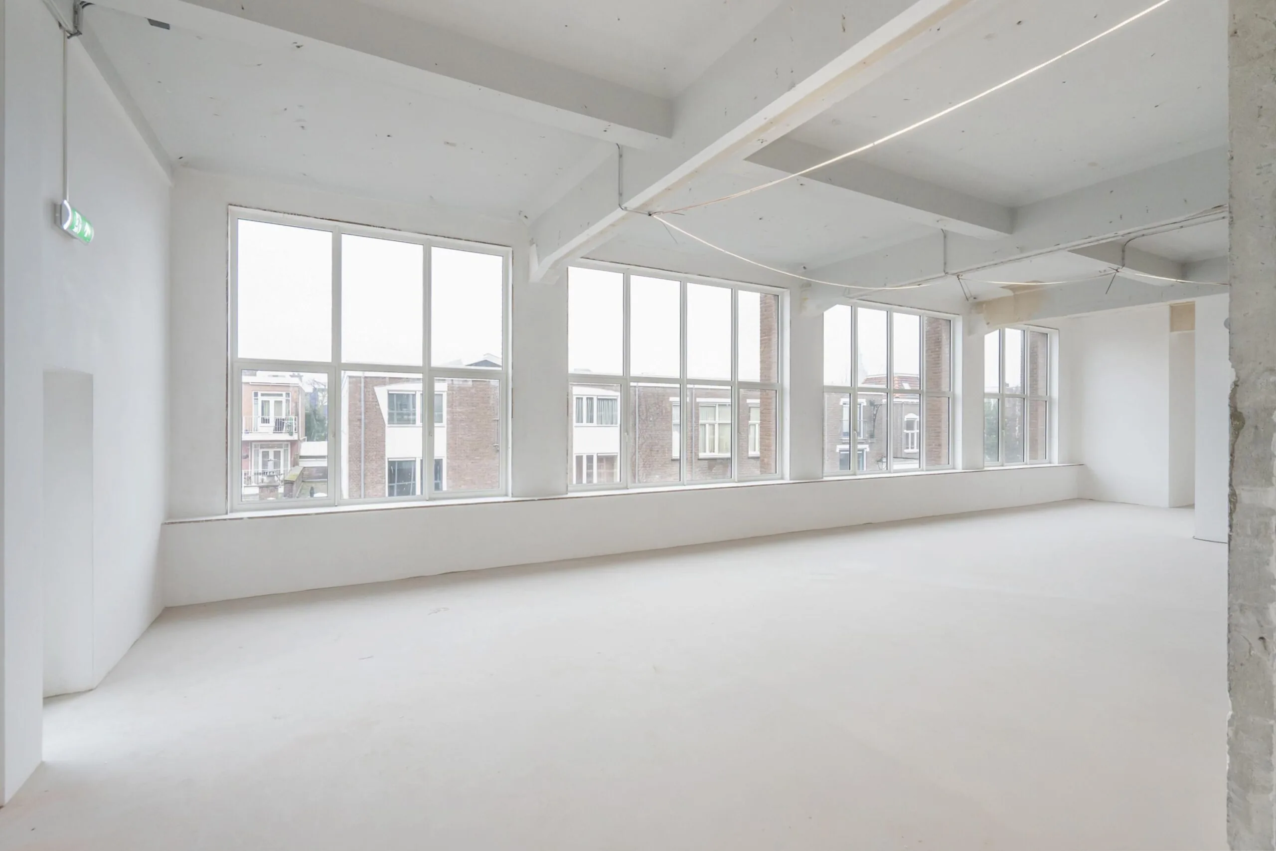 Een lege, lichte kamer met grote ramen die uitzicht bieden op nabijgelegen gebouwen.