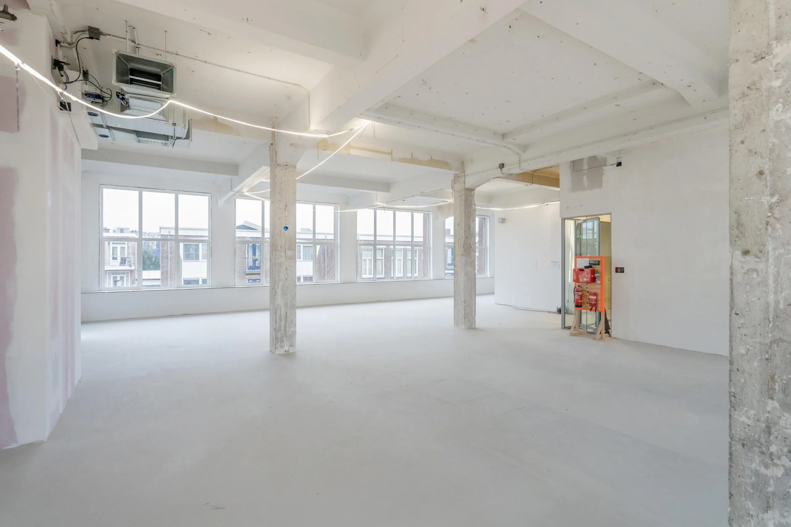 Een ruime, onafgewerkte industriële kamer met grote ramen en betonnen pilaren.