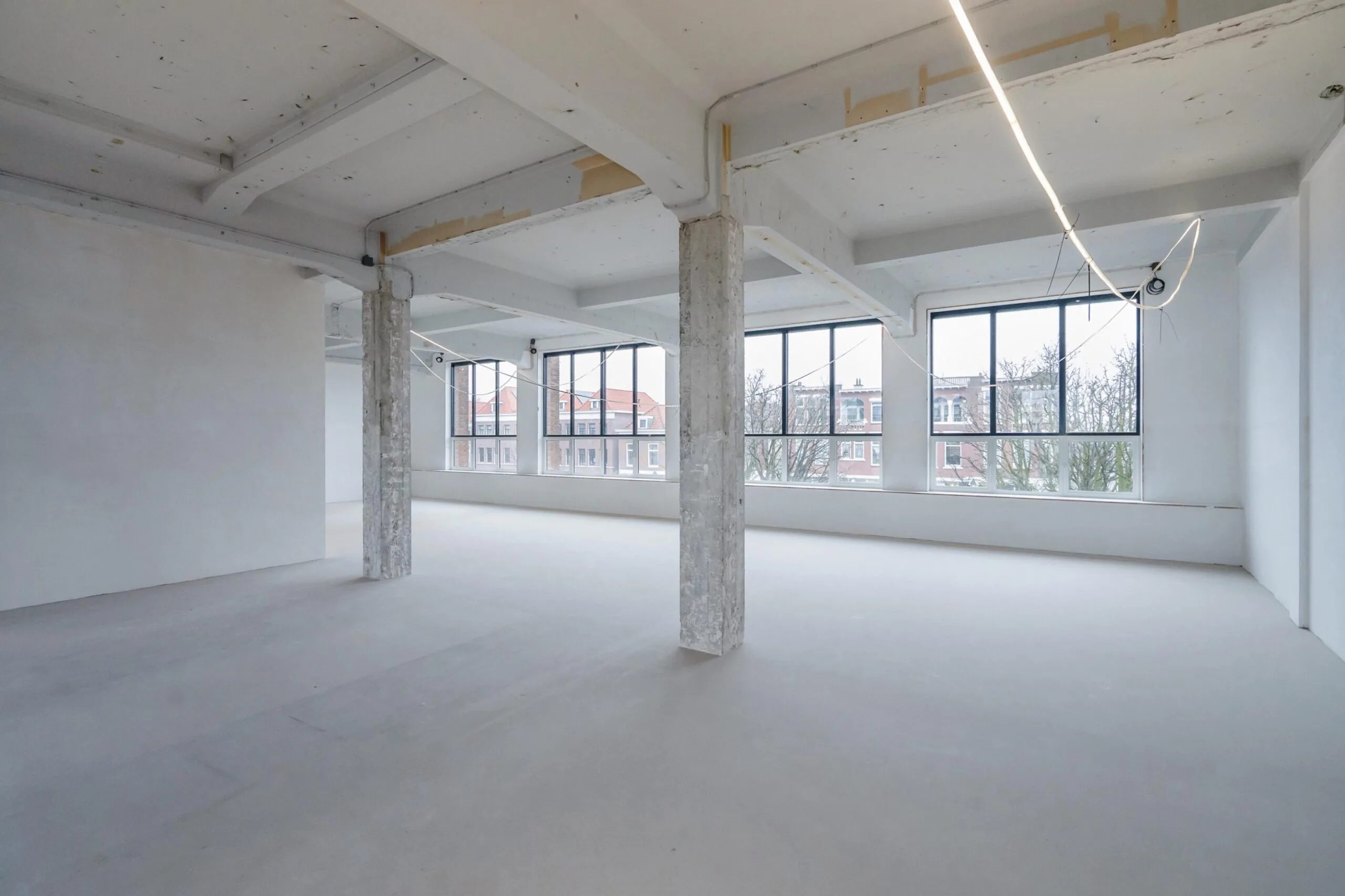 Lege industriële ruimte met betonnen pilaren en grote ramen die uitzicht bieden op omliggende gebouwen.