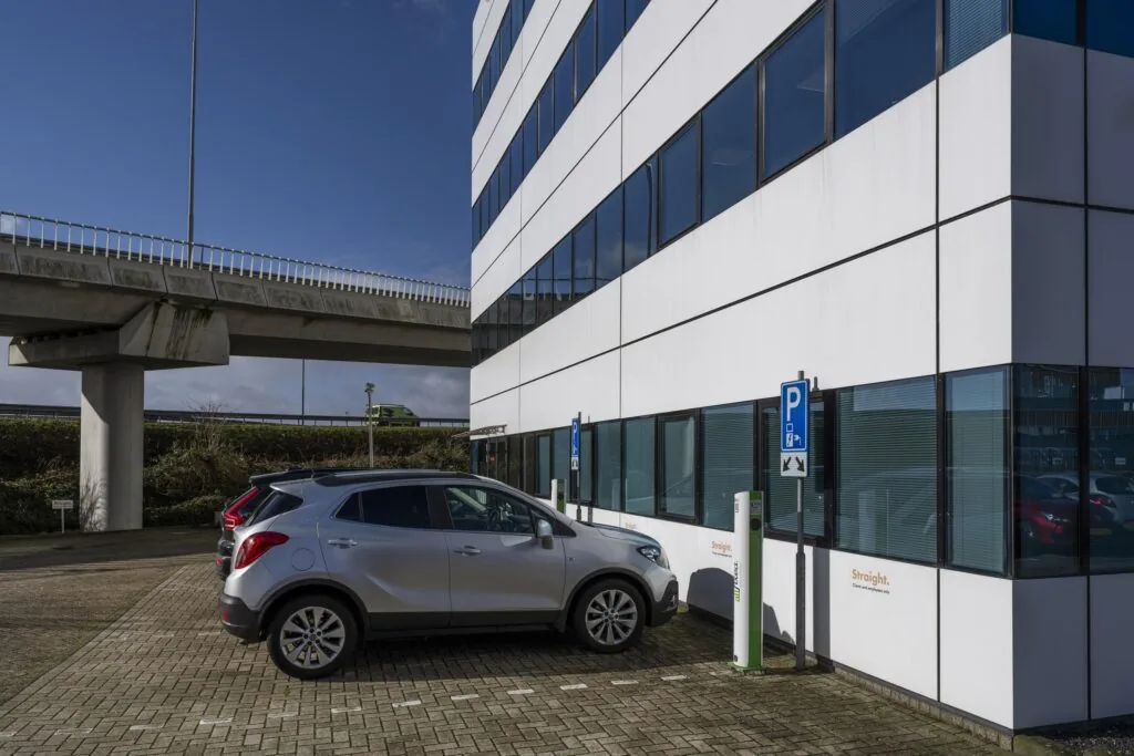 Een zilverkleurige auto is geparkeerd bij een modern kantoorgebouw naast een parkeerbord en laadpalen op de Kuiperbergweg.